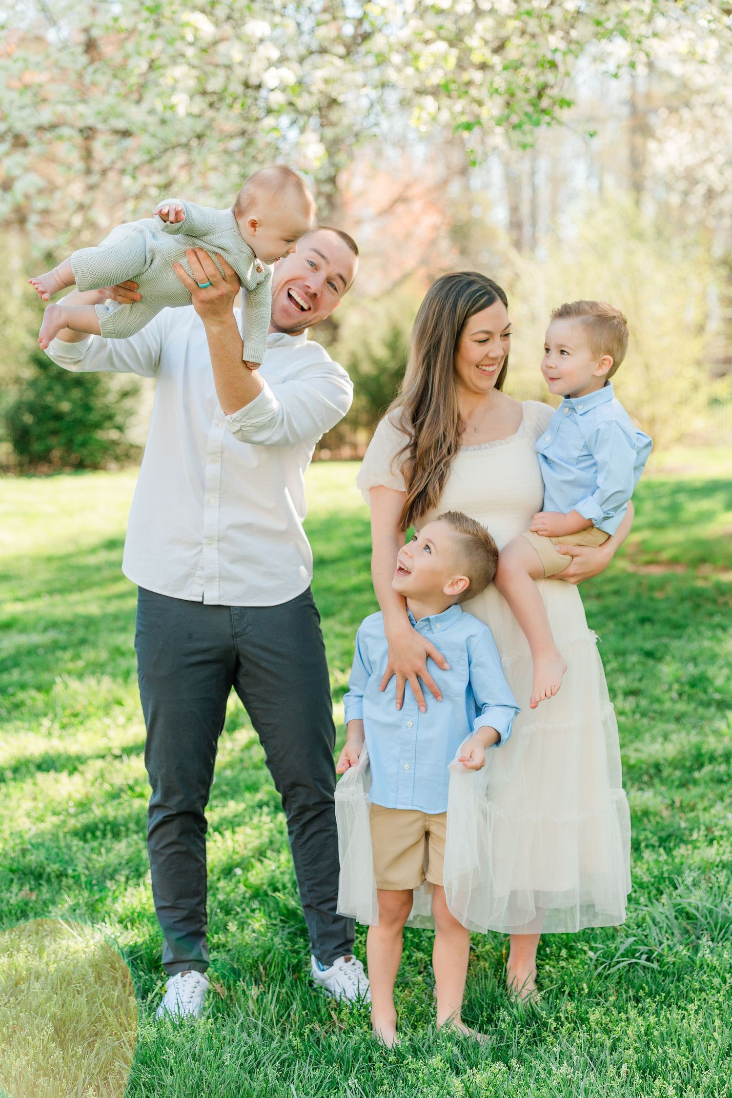 A joyful outdoor family photoshoot on green grass with everyone wearing coordinated white and light blue outfits.