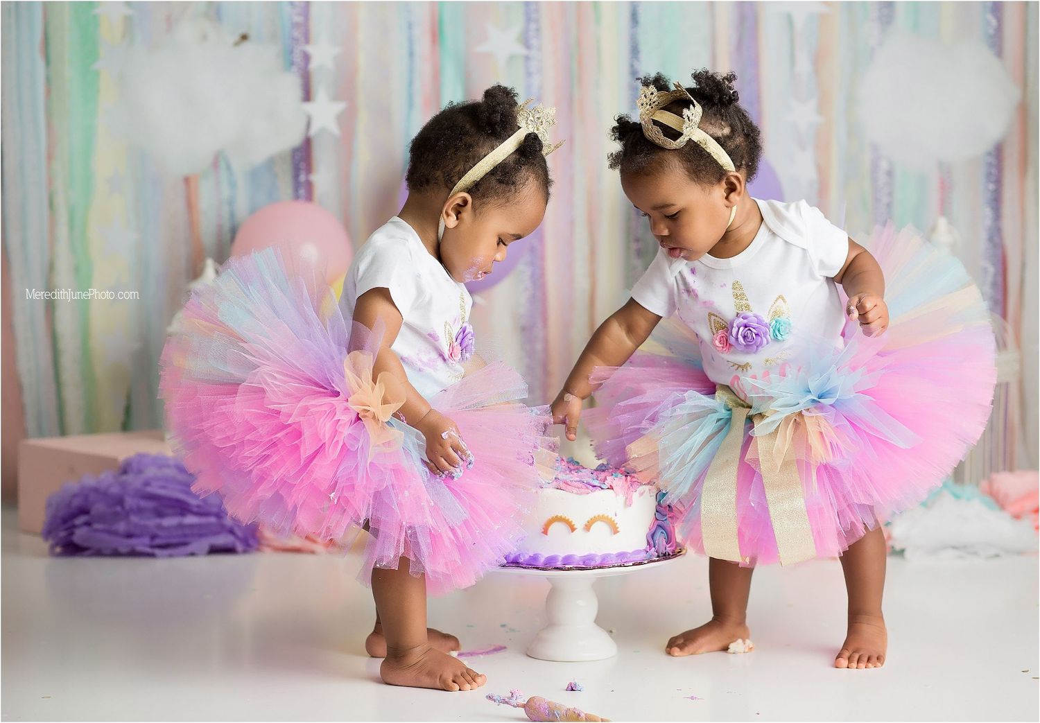 Two children in pastel tutus and tiaras sit together on a white floor with pink and purple decorations.