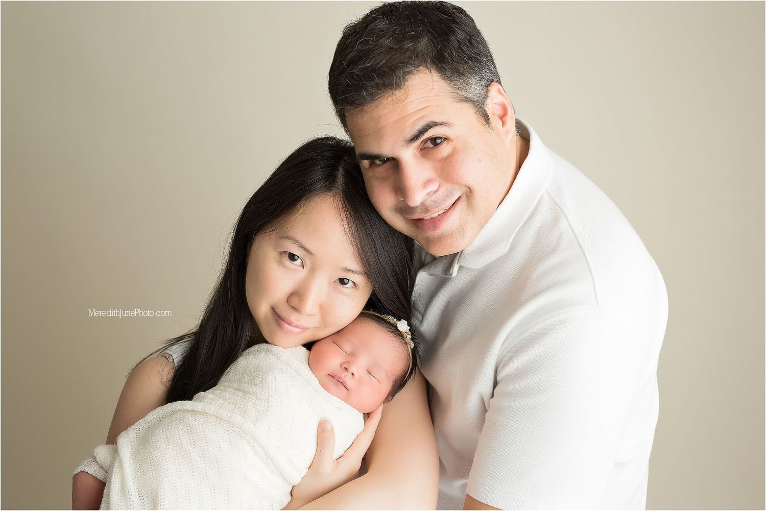 A couple in white clothing holds their newborn baby in a tender studio portrait.