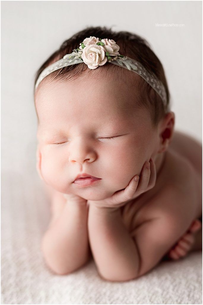 A sleeping newborn baby wearing a delicate floral headband lies peacefully on soft white fabric.