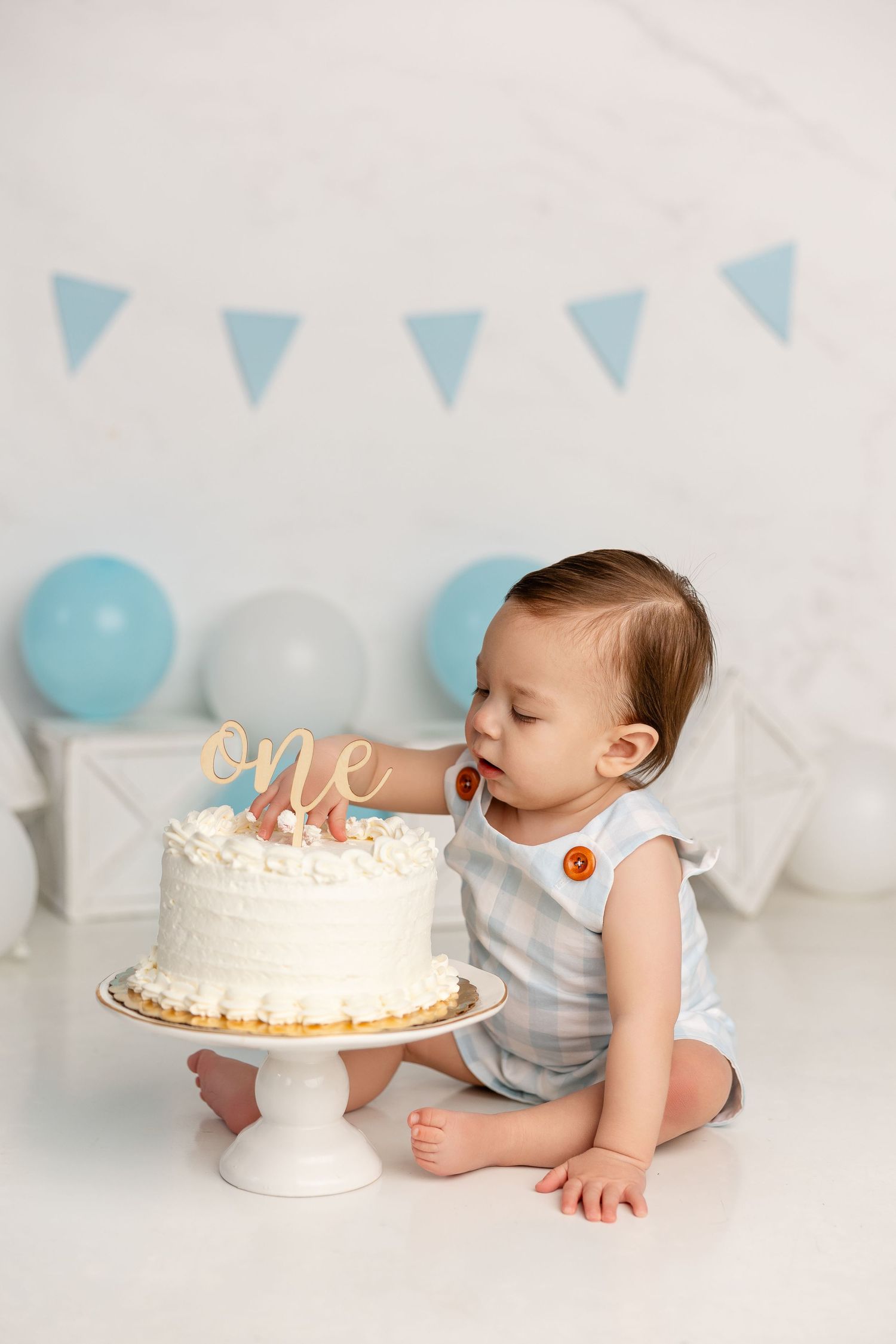 Baby in striped outfit reaches for white birthday cake with gold 'one' topper and blue party decorations.
