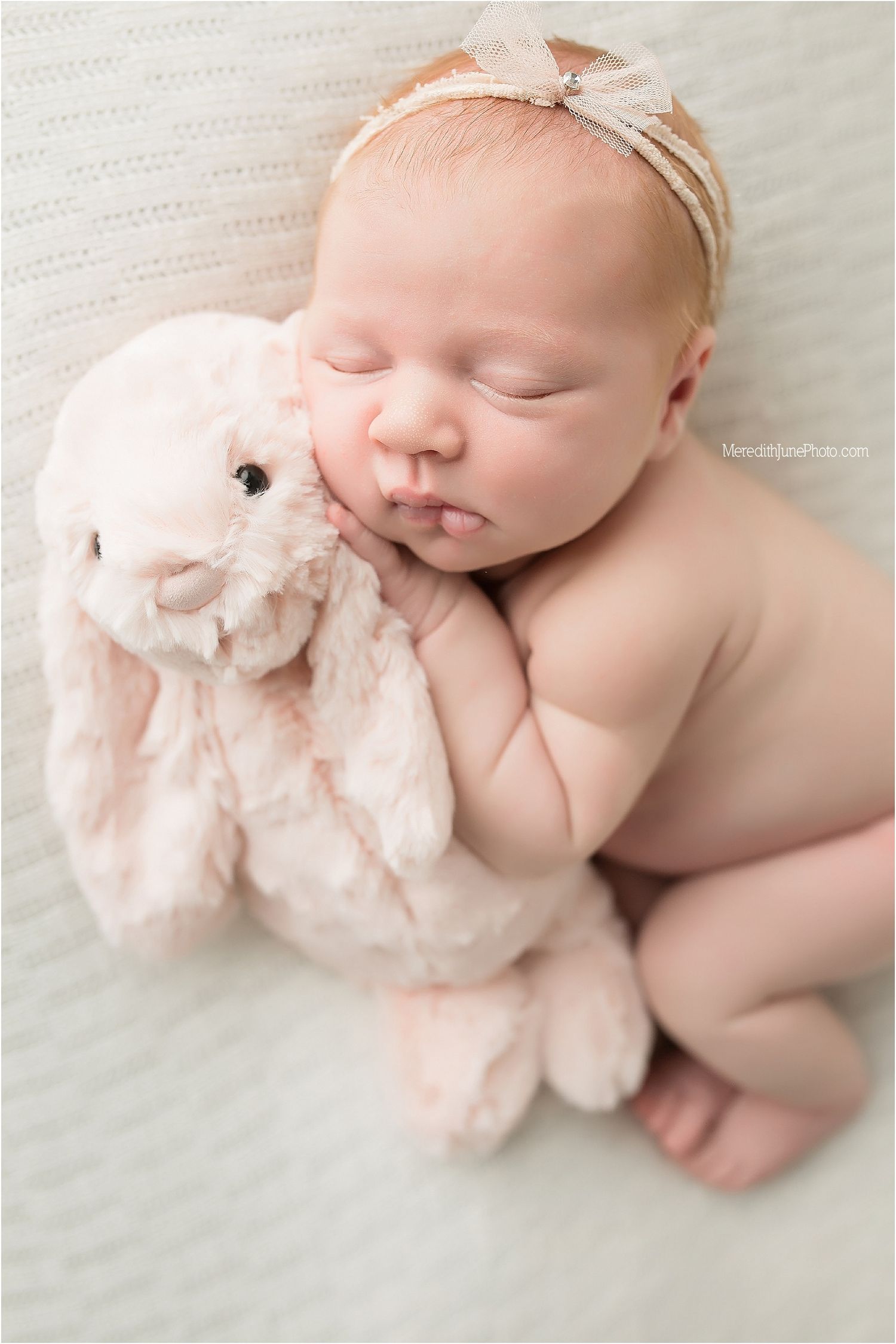 A sleeping newborn lies peacefully on white bedding while cuddling a soft pink plush bunny toy.