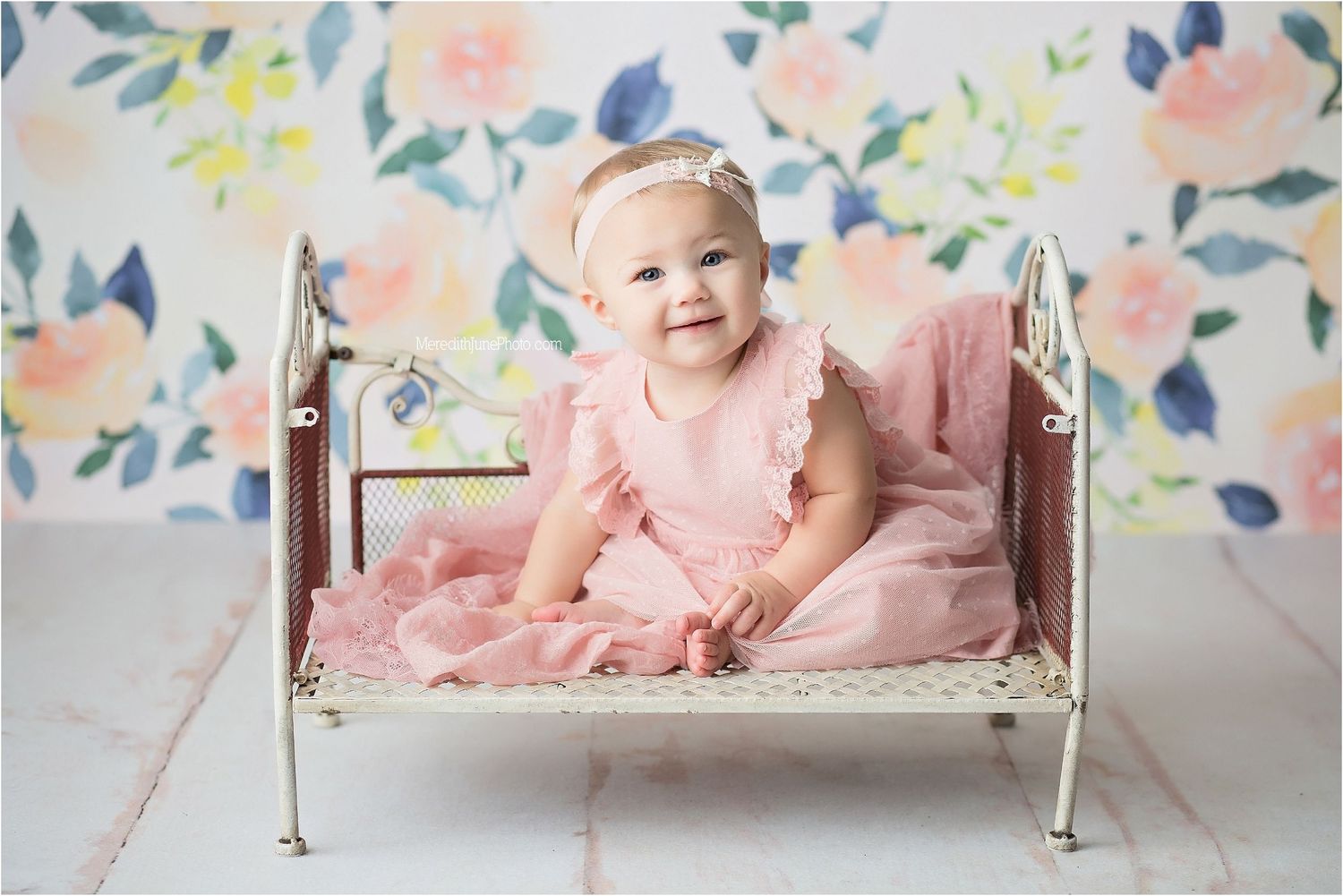 Baby sitting in vintage metal bed with pink dress against floral wallpaper backdrop.