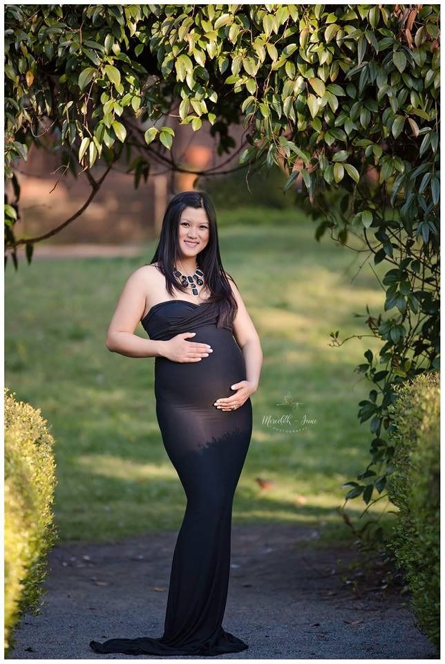 Maternity photo session in a garden setting with a black dress and natural sunlight filtering through tree leaves.