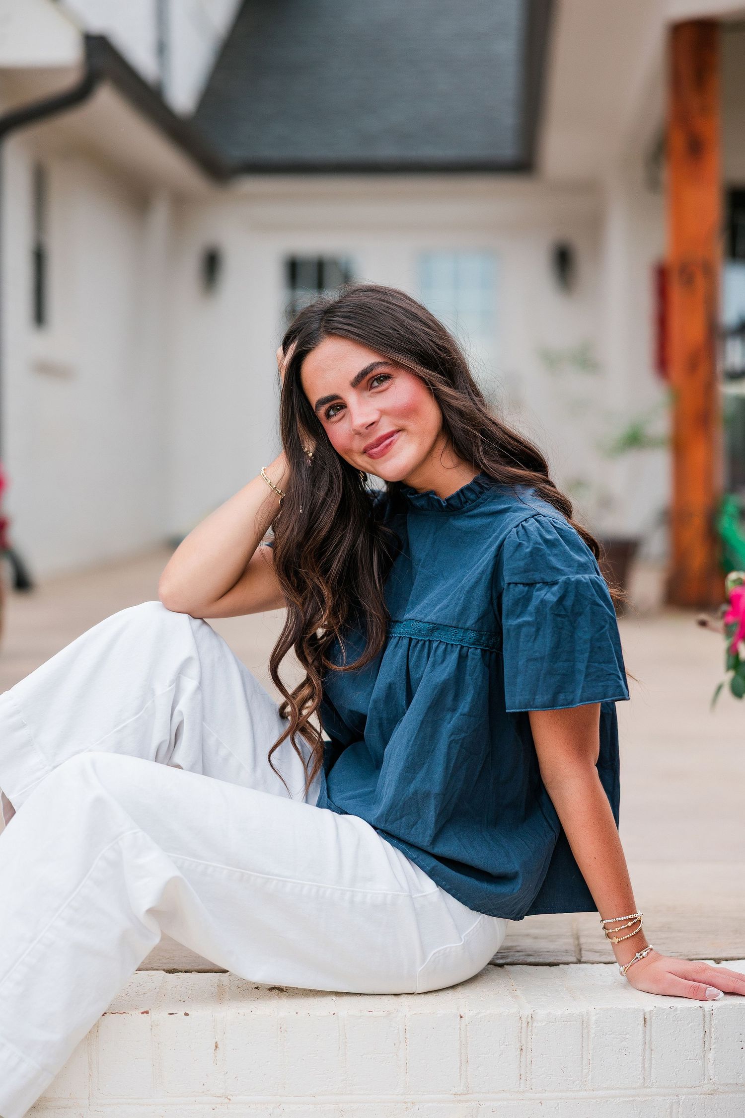 Woman in teal ruffled top and white pants sitting outdoors, smiling gracefully near a white building.