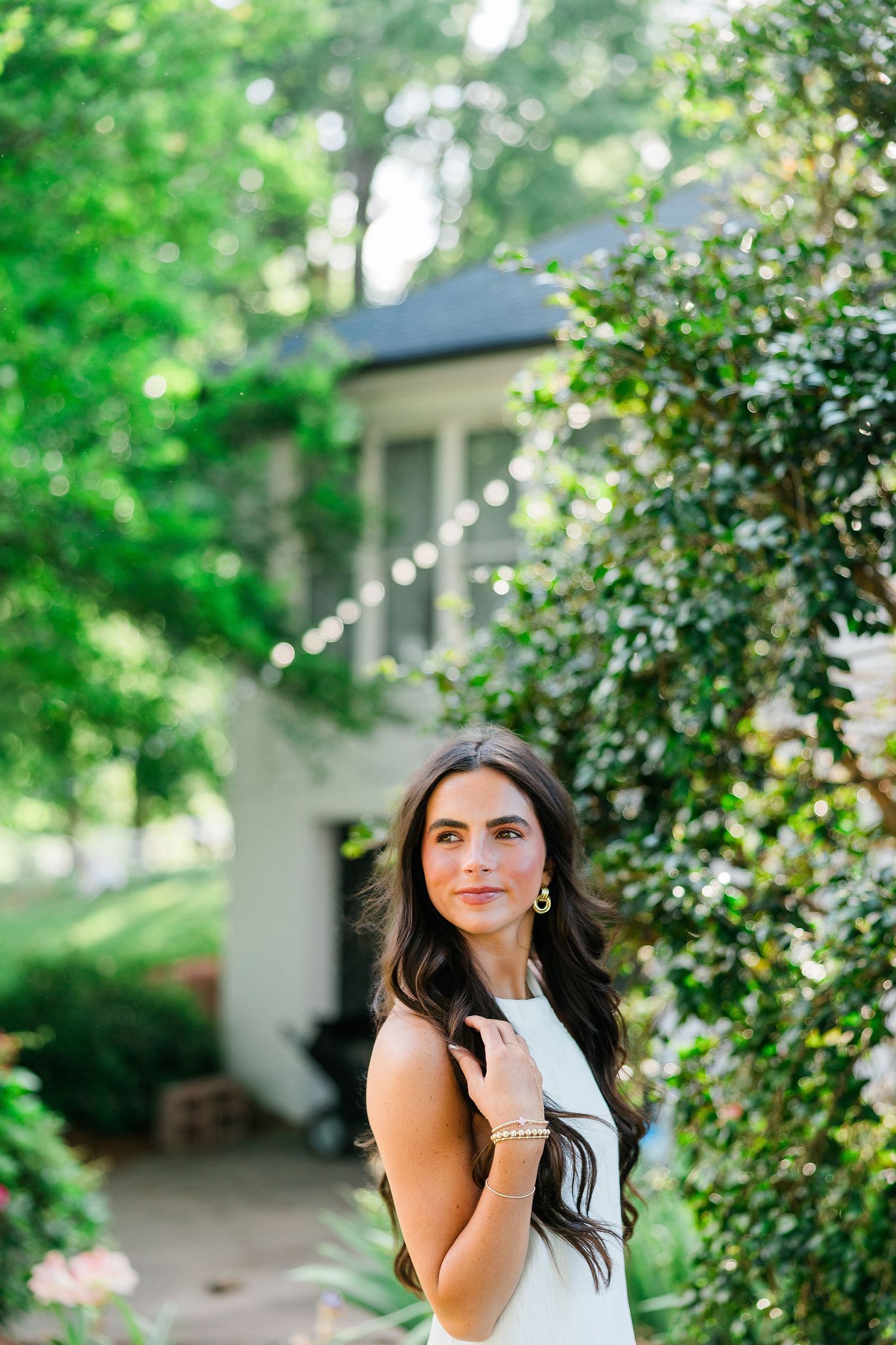A young woman in a white dress poses gracefully among lush green bushes near a white house.
