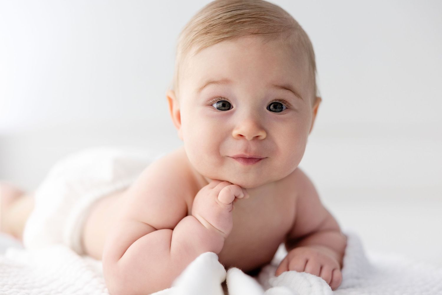 Baby in light-colored outfit lies on soft white surface with gentle expression.
