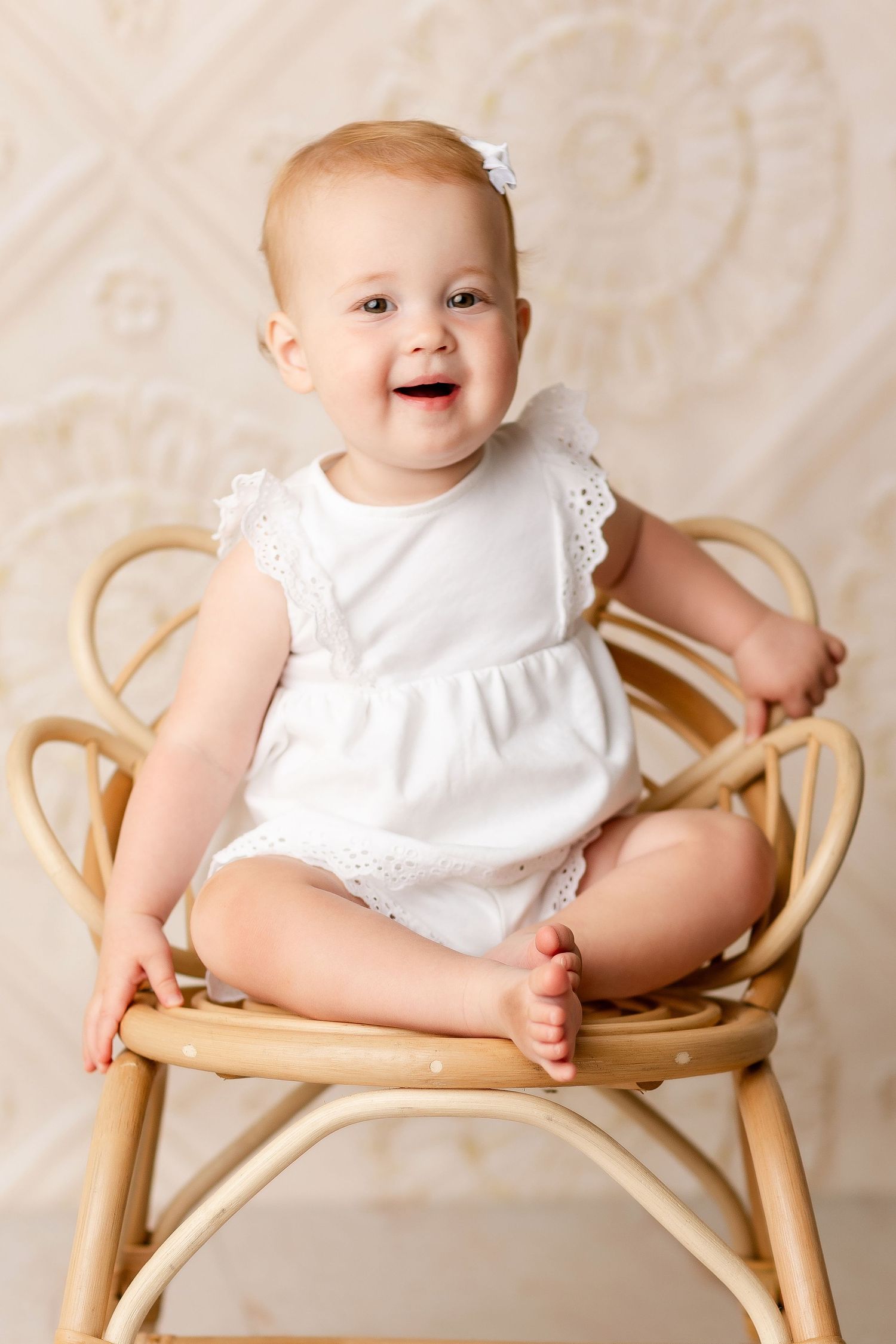 Baby in white dress sits and smiles on wooden chair during photo session.