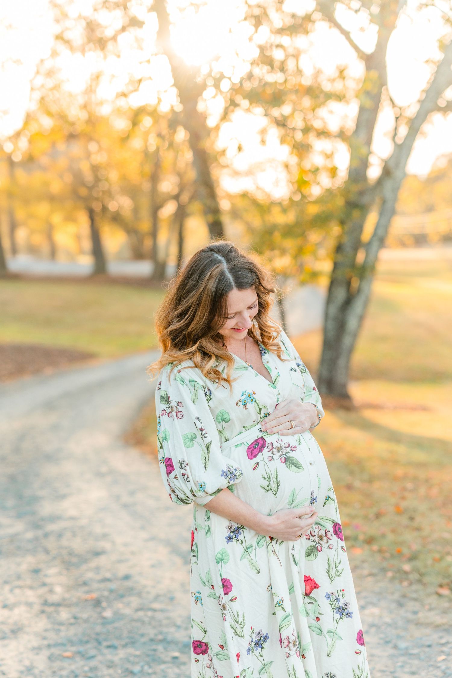 A pregnant woman in a floral dress stands on a path during golden hour with autumn trees in the background.