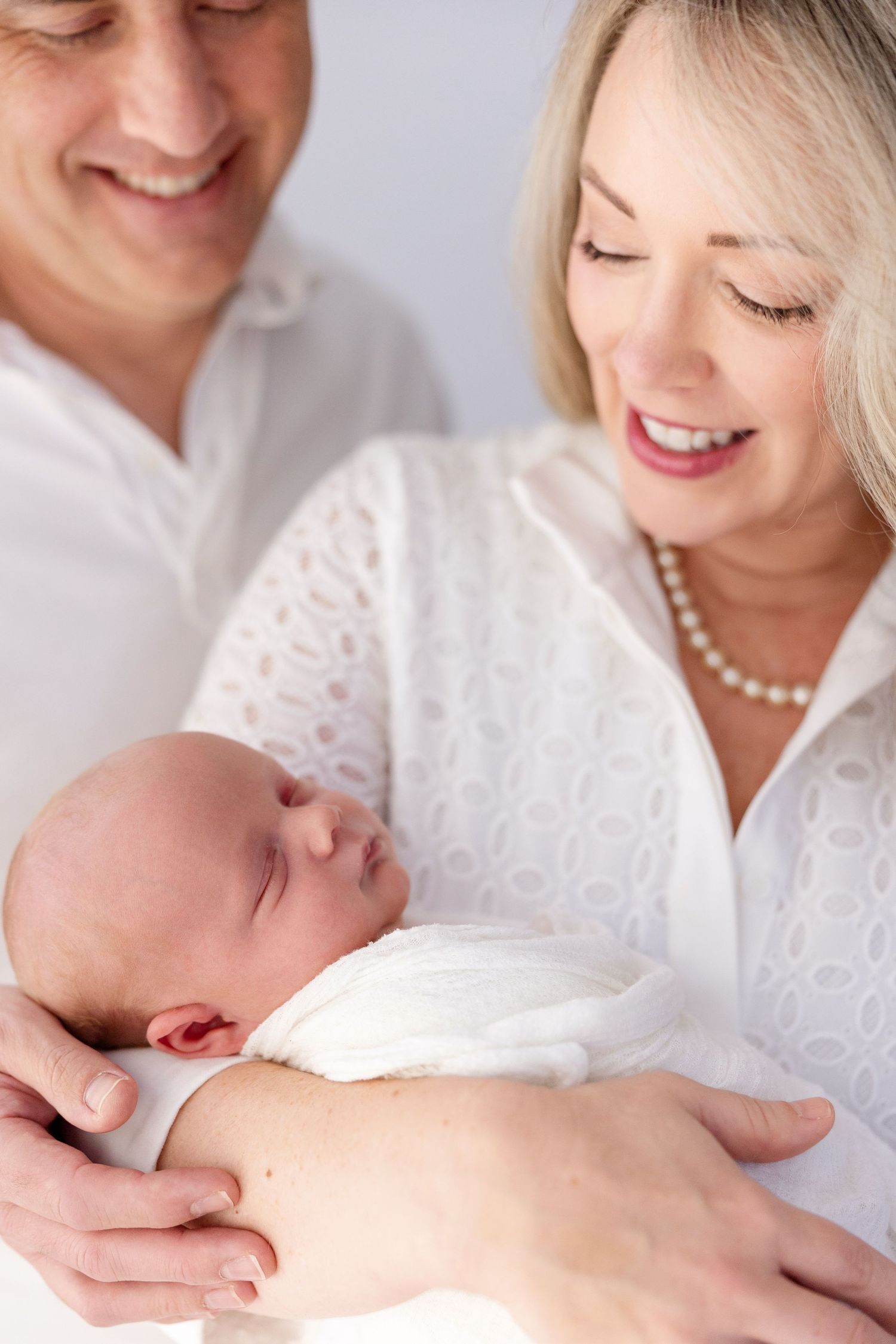 Smiling parents in white clothing lovingly hold their sleeping newborn baby wrapped in a white blanket.