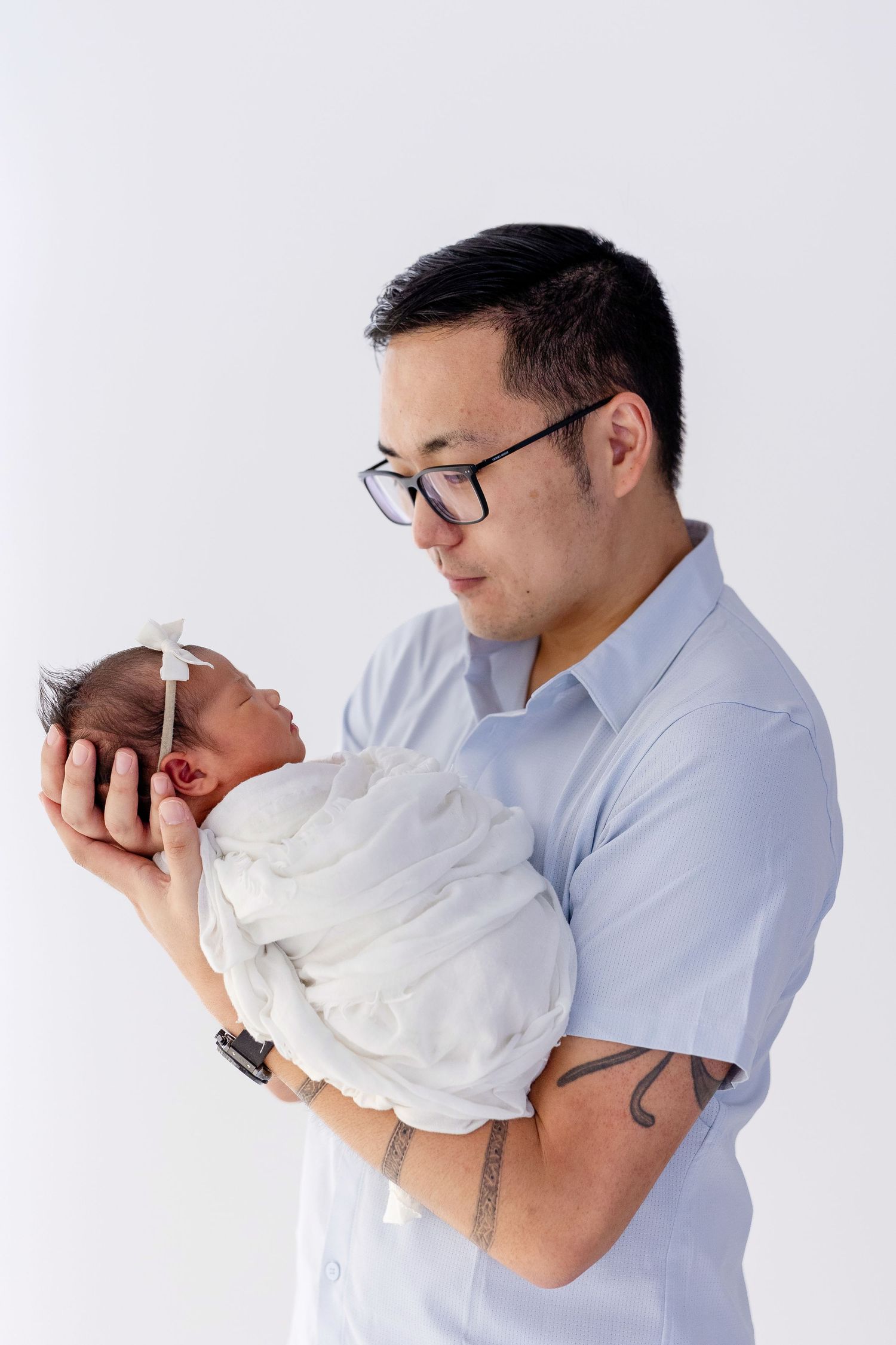 A man with glasses and tattoos tenderly holds a swaddled newborn baby girl with a white bow against a white background.