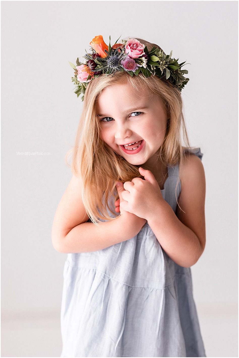 Child wearing floral crown and gray dress laughs joyfully against white backdrop.