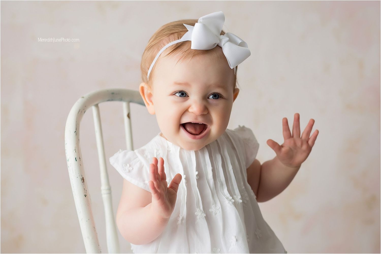 A joyful baby in a white dress and bow sits in a vintage white chair against a neutral backdrop.