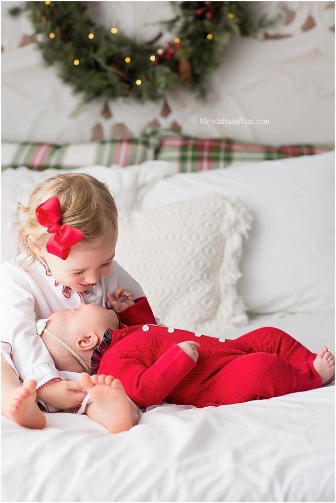 A baby in red holiday pajamas lies on white bedding beneath Christmas decorations.