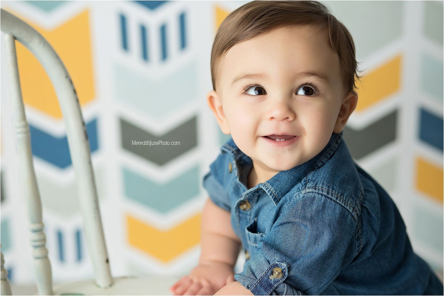 Baby wearing denim shirt smiles and poses against chevron patterned wall background.