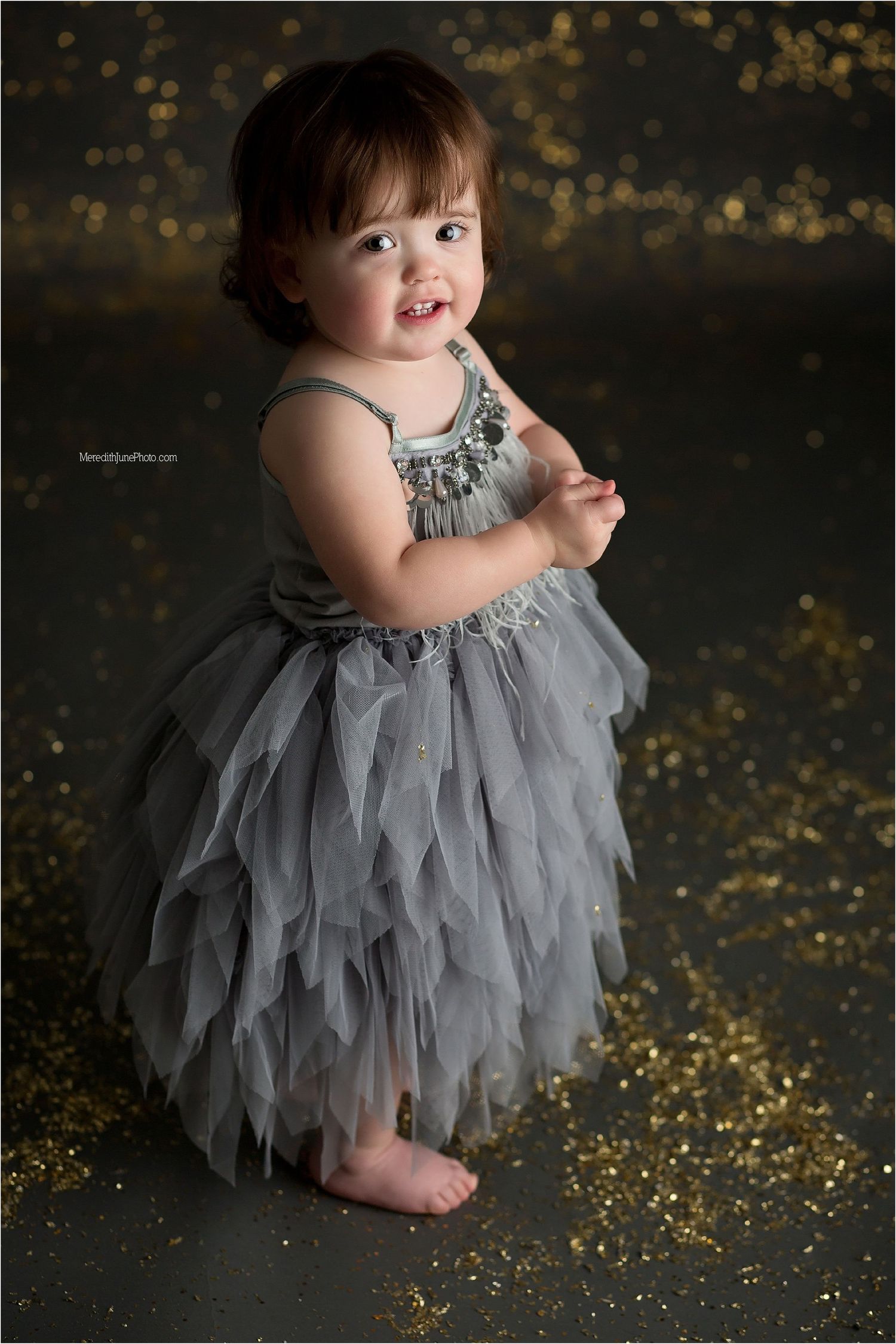 A silver and grey tulle dress twirls against a dark backdrop with scattered gold glitter on the floor.
