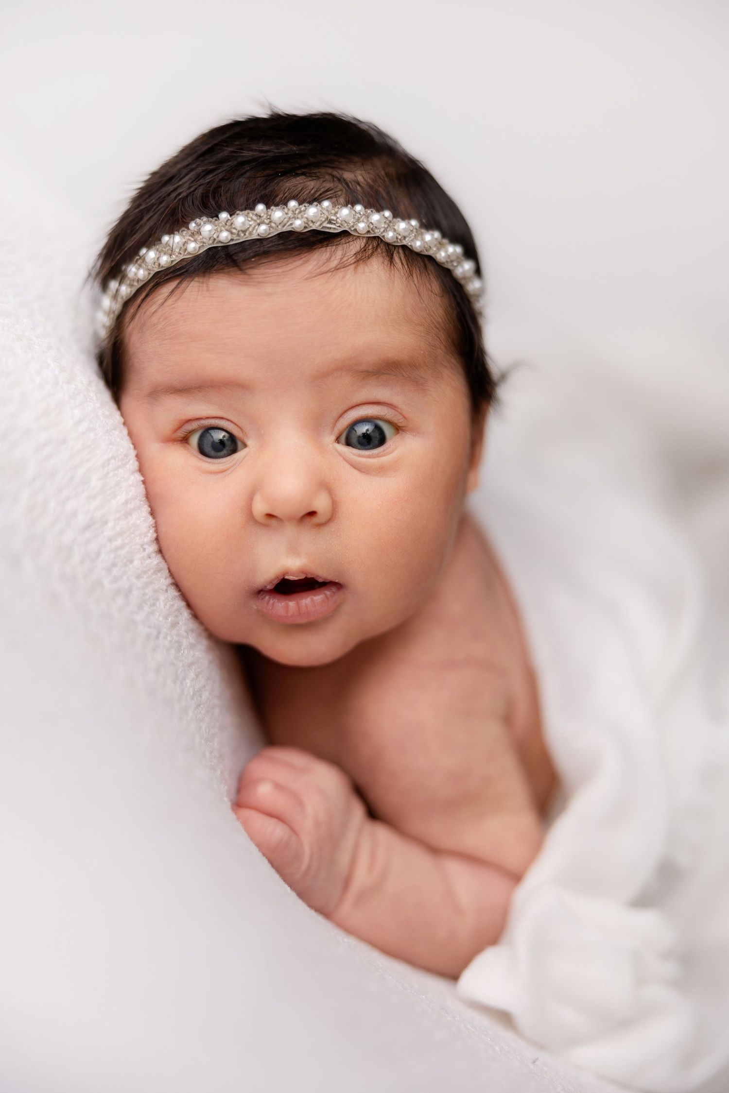 Sweet baby wearing delicate crystal headband peeks out from soft white blanket with wide-eyed expression.
