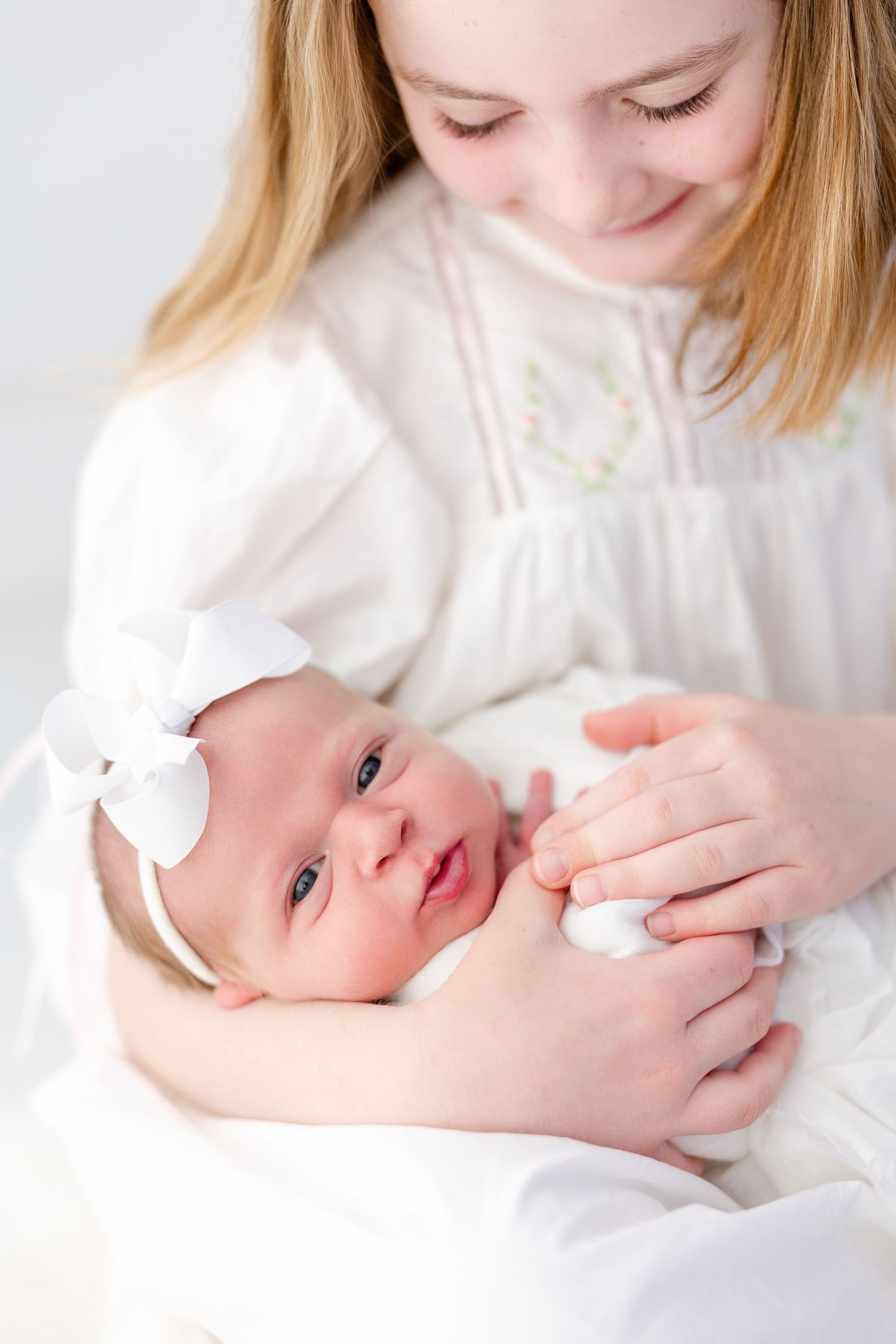 A newborn baby wearing a white bow headband is held lovingly in white clothing against a light background.