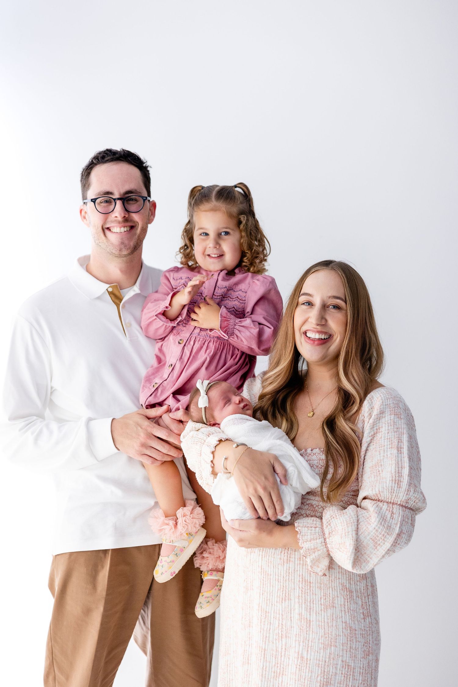 A family wearing coordinated outfits in white and tan poses together against a white backdrop for studio portraits.