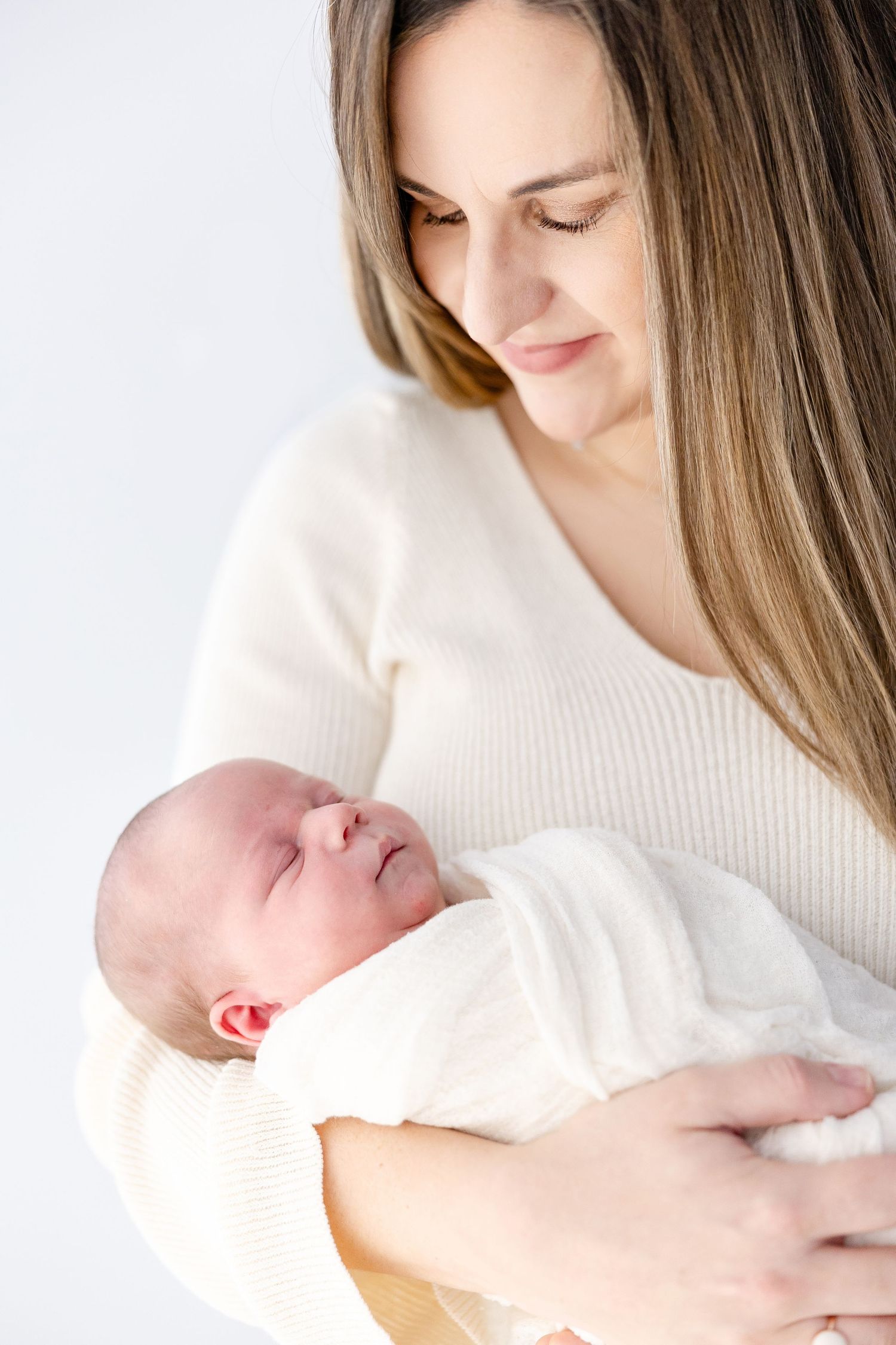A tender moment captured as a mother in a cream sweater holds and gazes lovingly at her sleeping newborn baby wrapped in white.