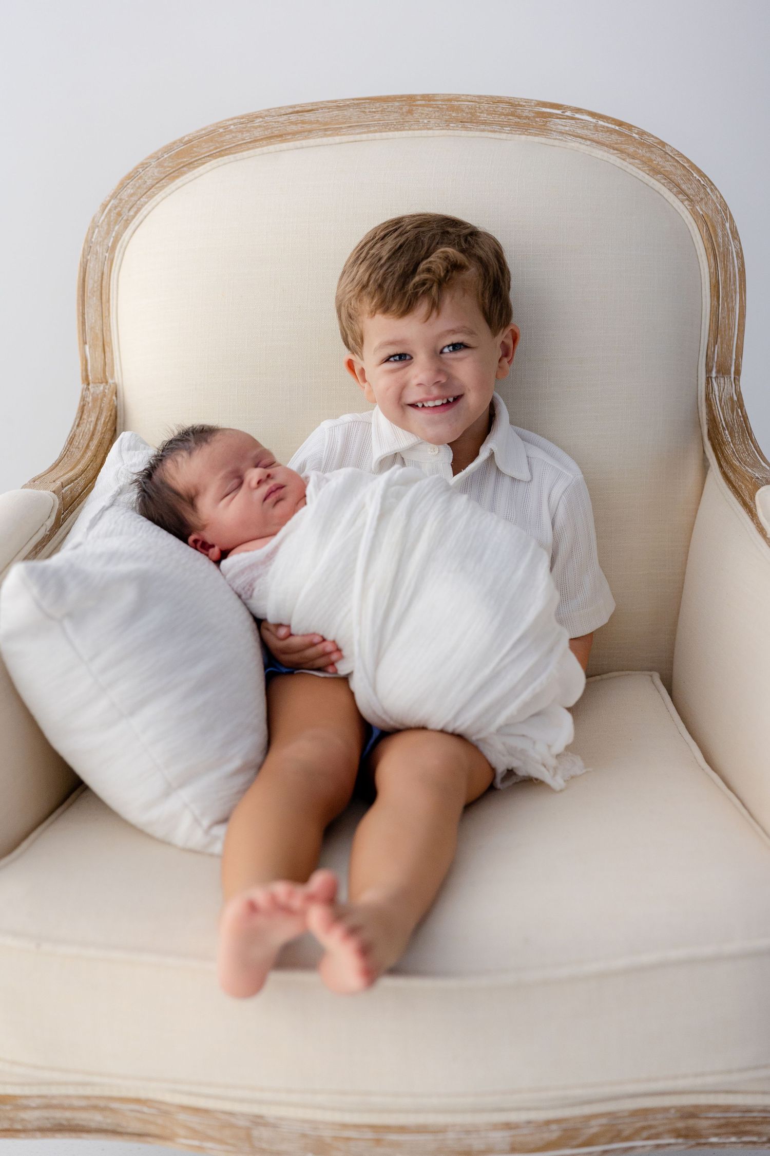 Smiling young boy in white shirt holds sleeping newborn baby sibling in cream armchair for a professional portrait.
