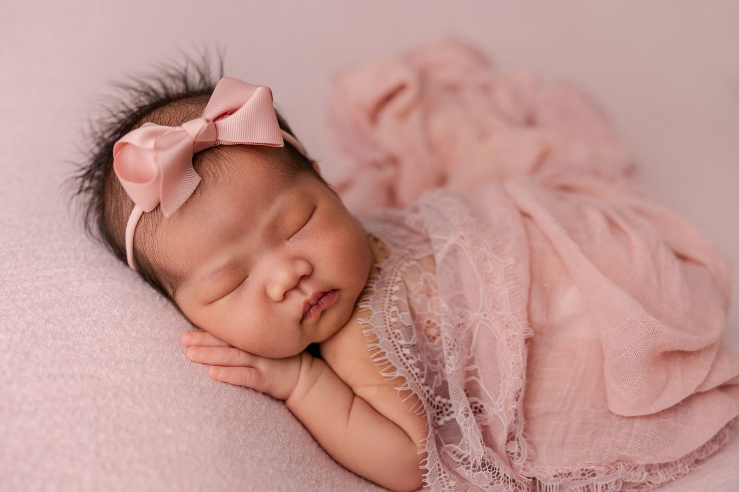 Newborn baby girl sleeping peacefully wrapped in pink lace with a pink bow headband.