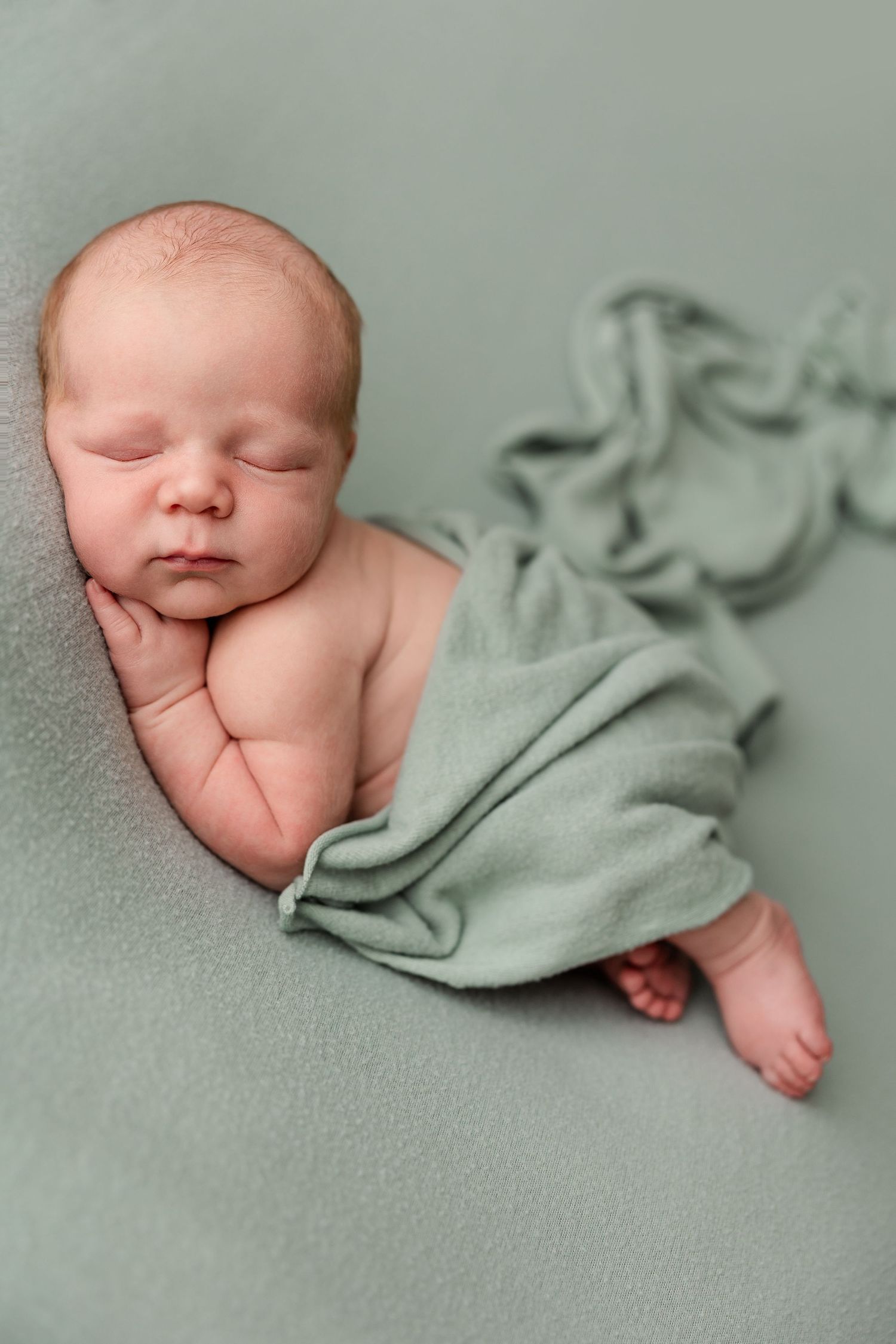 Newborn baby sleeping peacefully on a soft sage green blanket, gently wrapped in a matching green swaddle cloth.