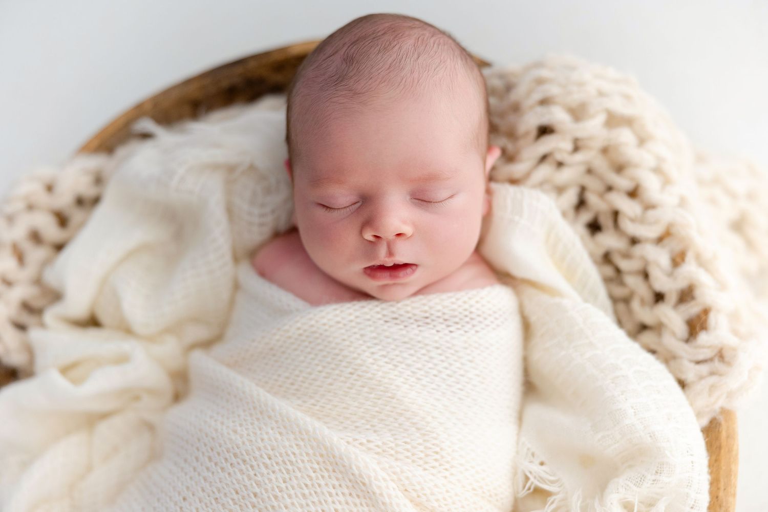 Newborn baby sleeping peacefully swaddled in cream knit wrap inside a wooden bowl with chunky knit blanket.