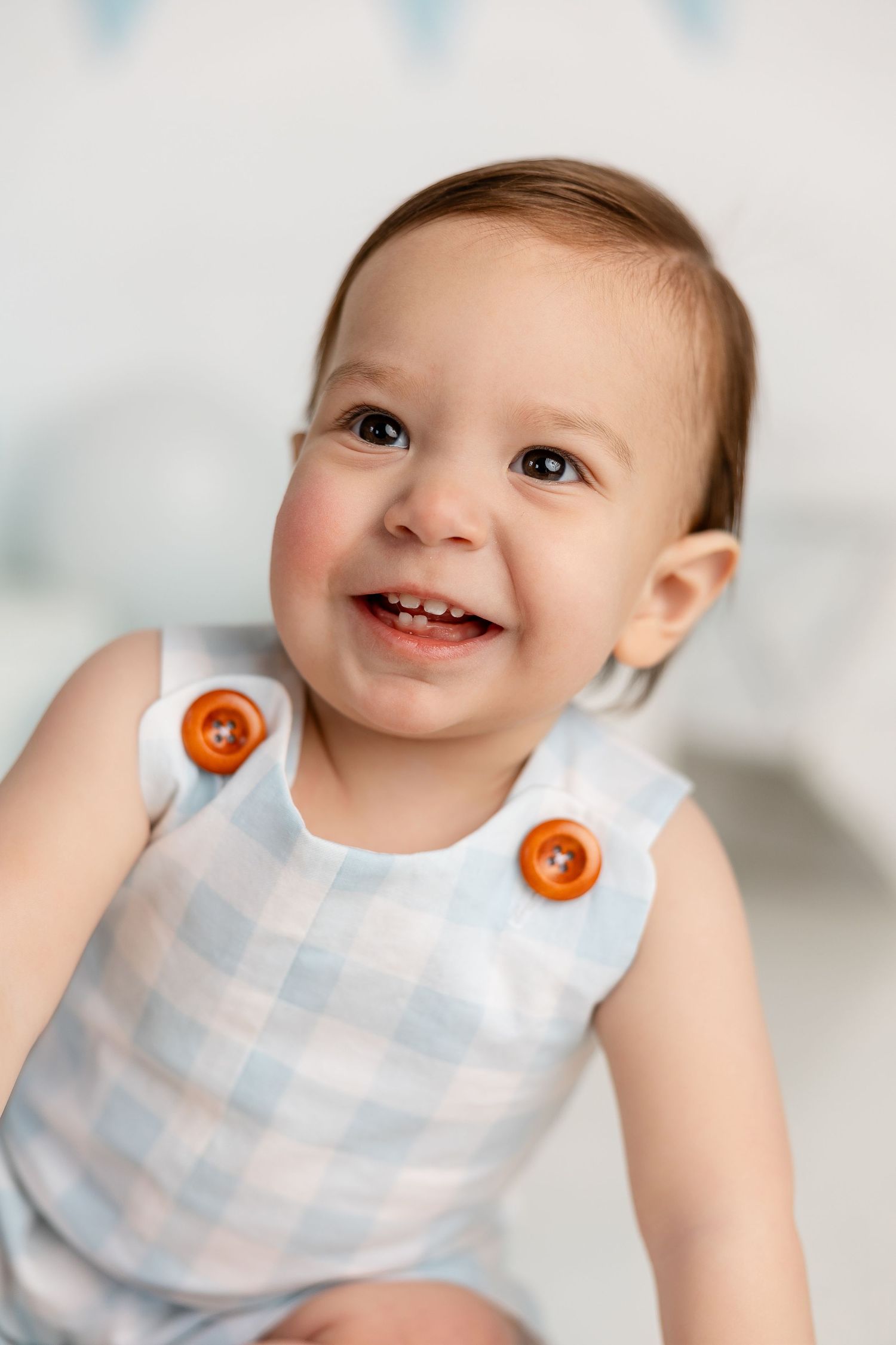 Smiling baby wearing a light blue checkered romper with orange buttons against a white background.