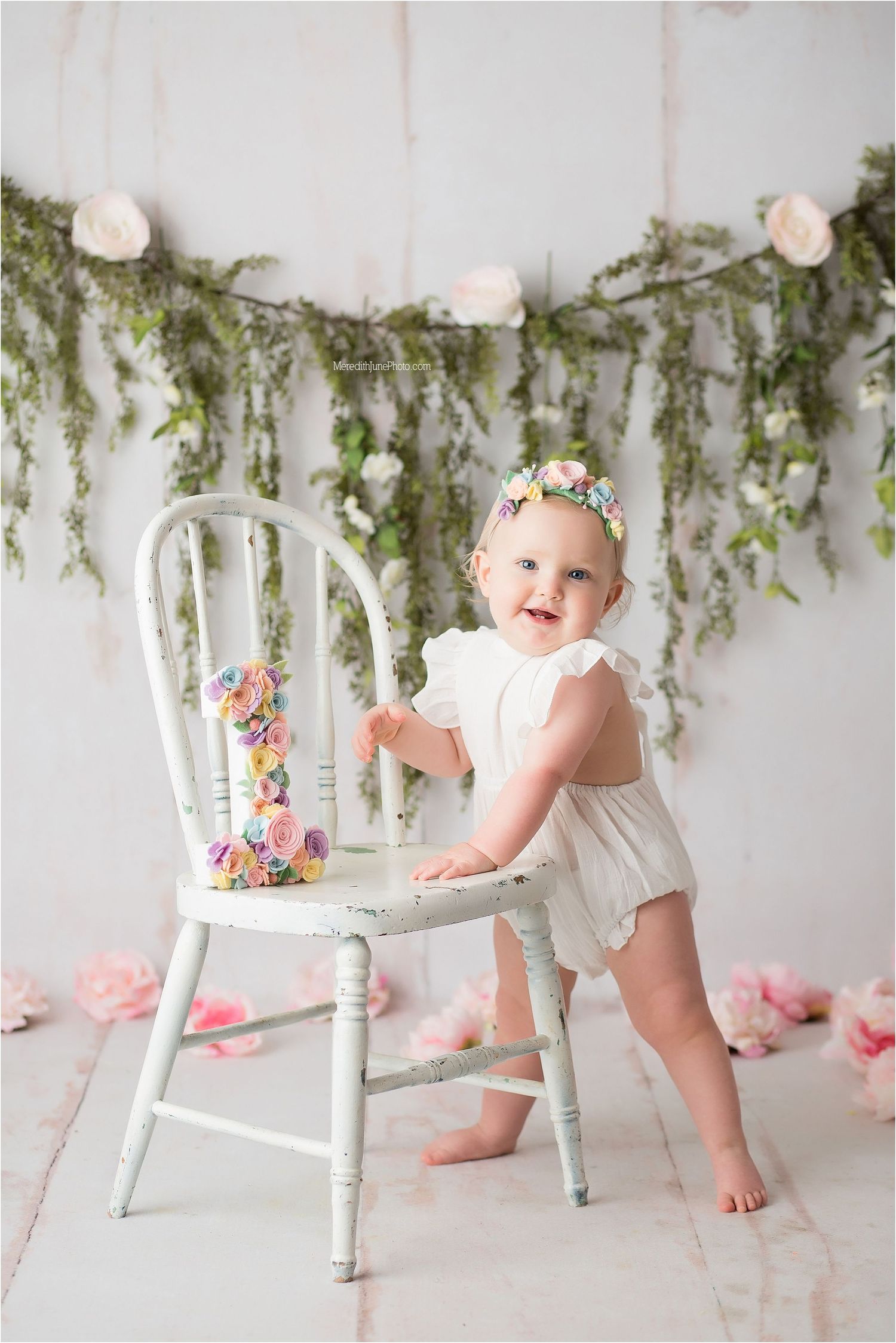 Baby in white dress plays near vintage white chair with floral decorations and greenery garland backdrop.