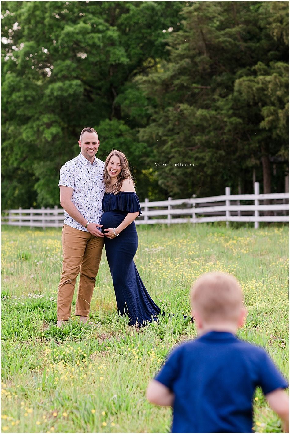 A couple shares a tender moment in a grassy field with white fence and wildflowers in the background.