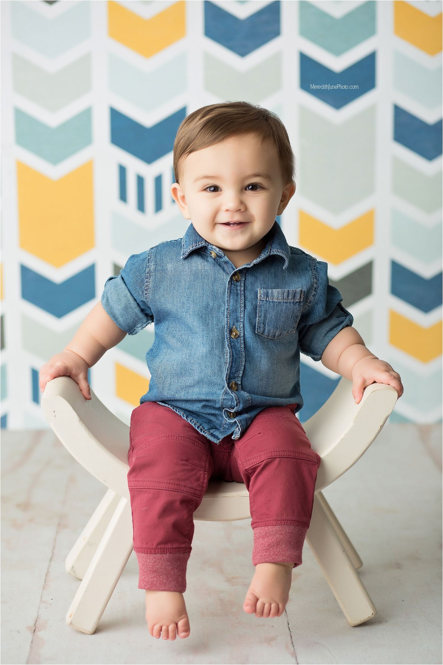 Baby in denim shirt and burgundy pants sits on white chair against chevron patterned wall.