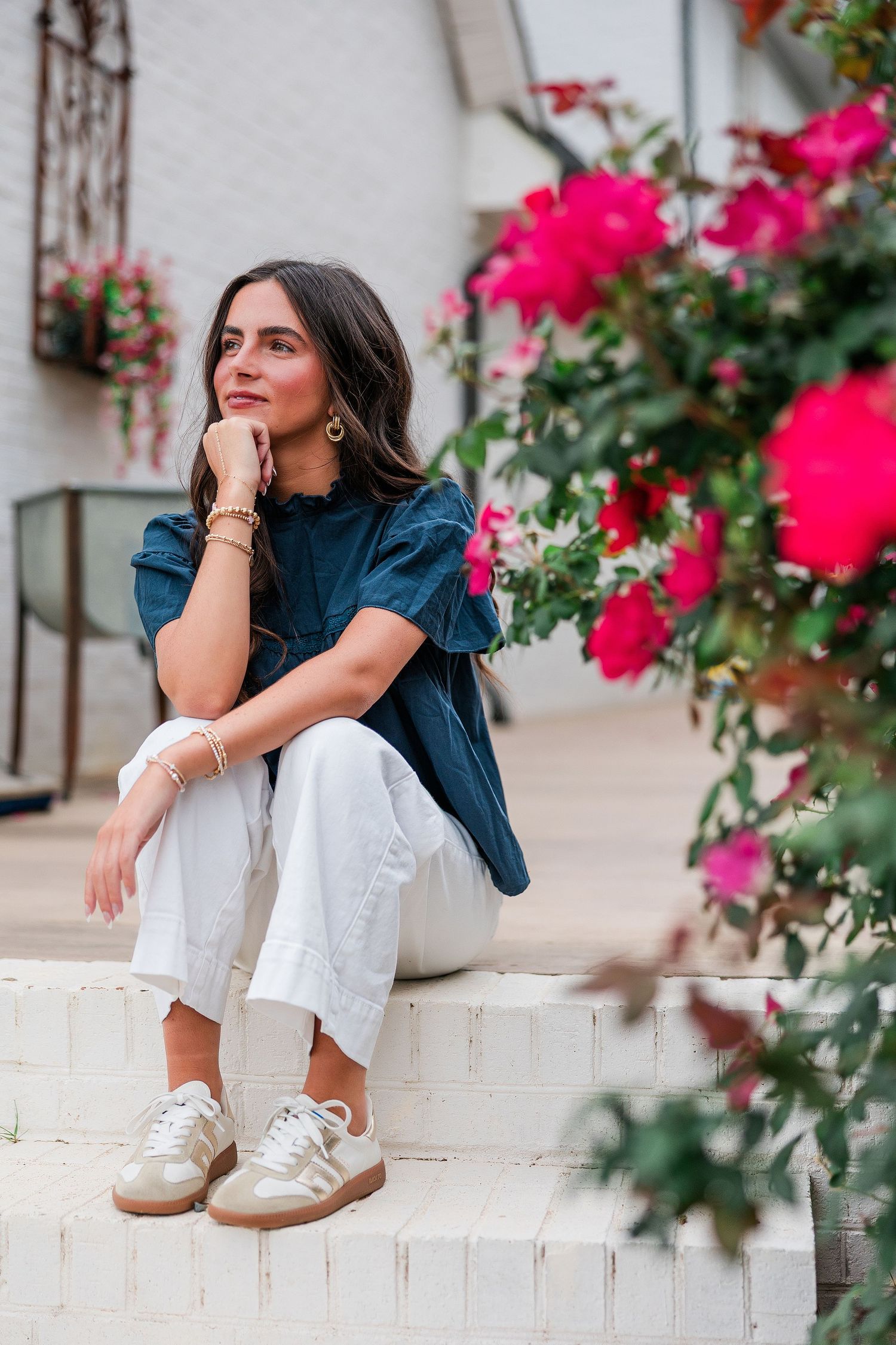 Woman in blue top and white pants sitting on steps surrounded by vibrant pink roses.