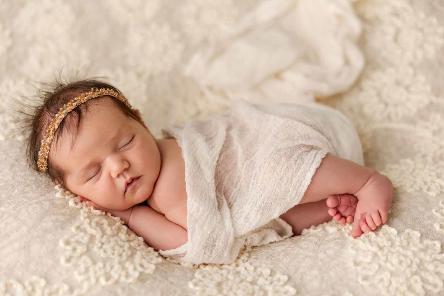 Sleeping newborn baby girl with a gold beaded headband wrapped in white gauze, lying on cream lace fabric in a newborn photoshoot.