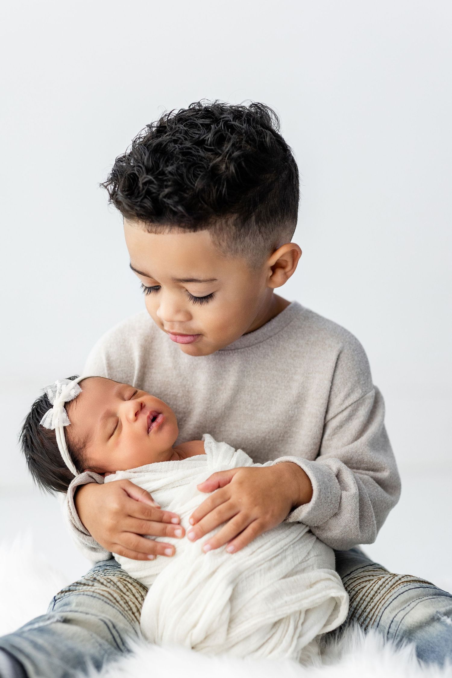Young child in gray sweatshirt tenderly holds and kisses newborn baby on white background.