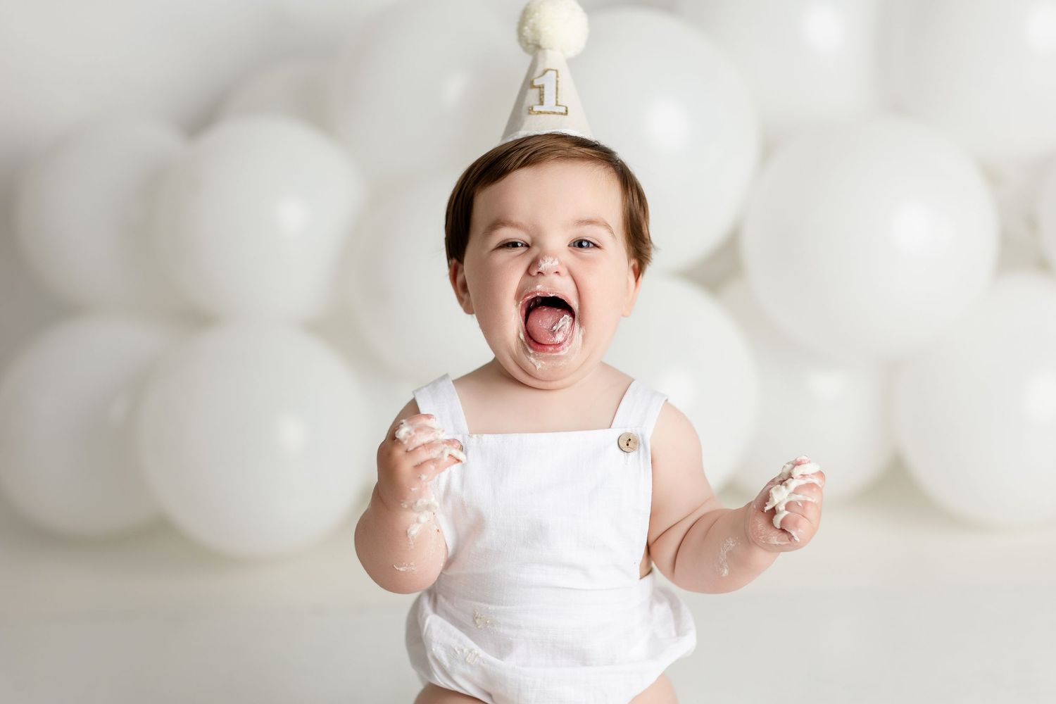Baby in white outfit against white balloon background during milestone photo session.