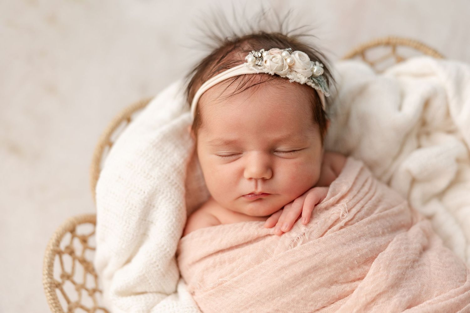Sleeping newborn baby girl in a pink swaddle wrap and floral headband resting in a cozy woven basket with a soft cream blanket.