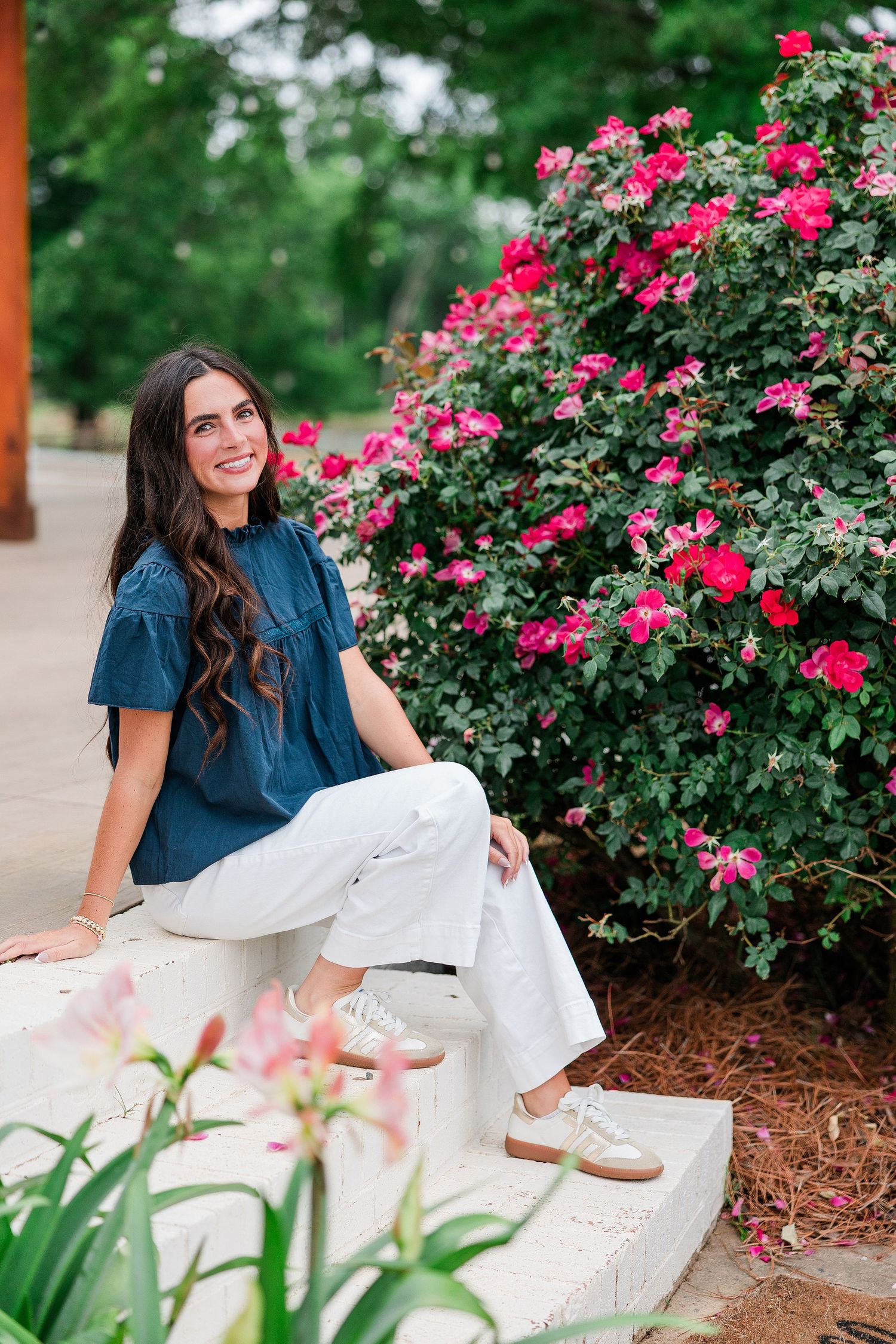 Young woman in blue top and white pants sitting near vibrant pink rose bushes in a lush garden setting.