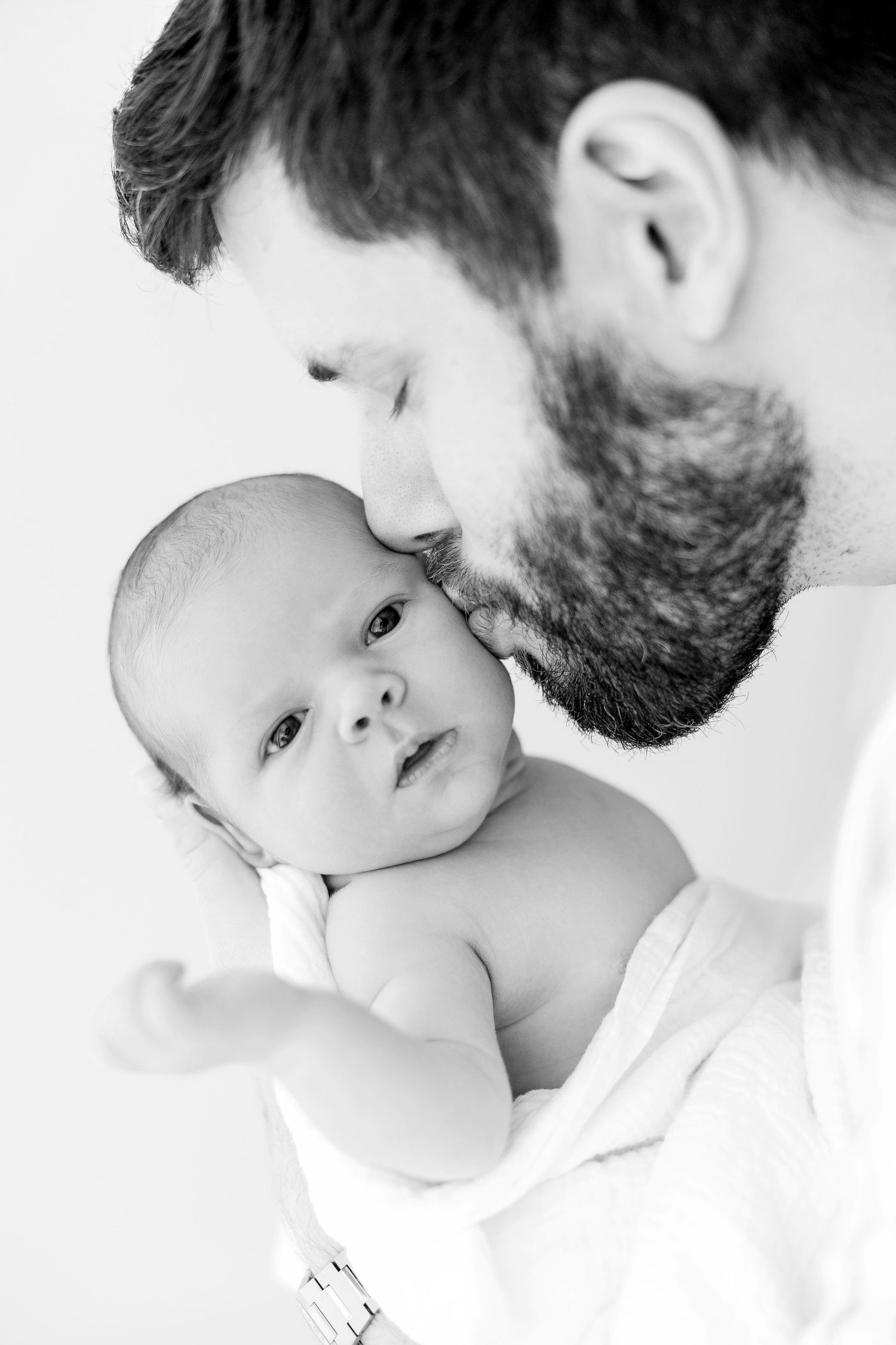 Black and white photo of a bearded father kissing his newborn baby tenderly.