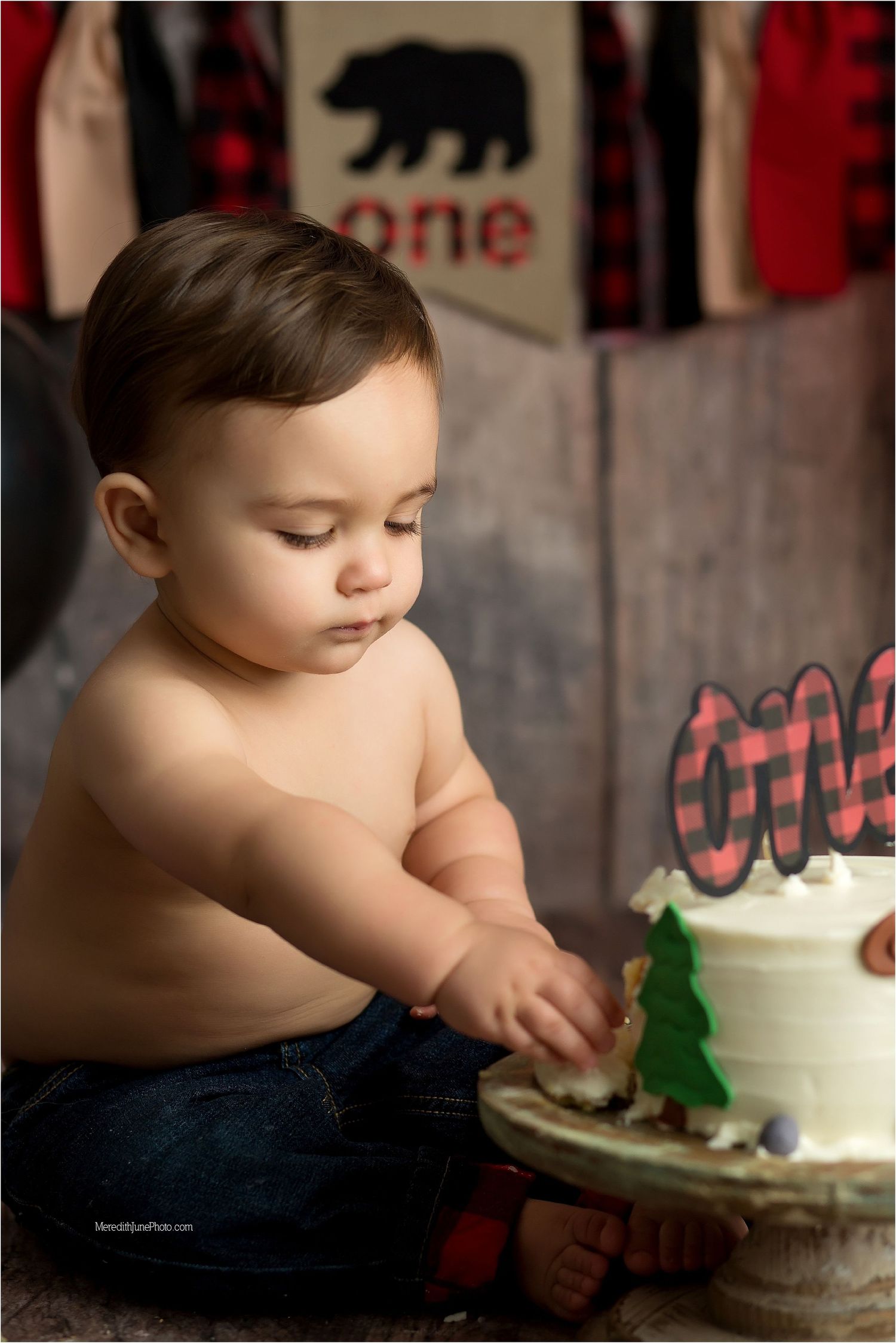 A small child in denim reaching for a white cake decorated with a green tree on a rustic wooden cake stand.