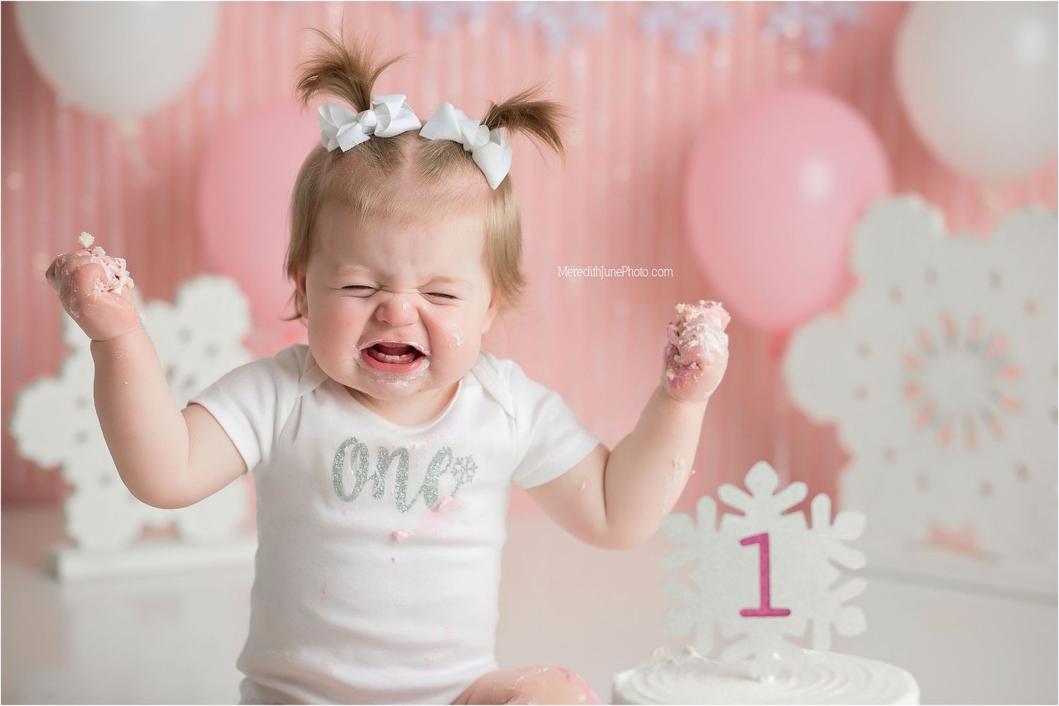 Baby in white shirt celebrates with cake during first birthday photoshoot with pink balloon backdrop.