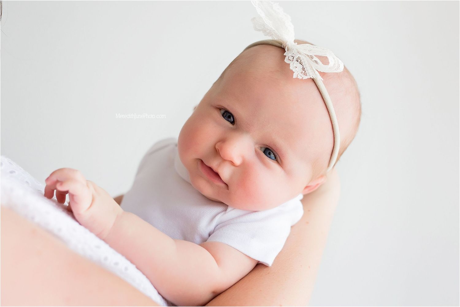 Baby wearing white onesie and lace headband lies on soft surface while smiling.