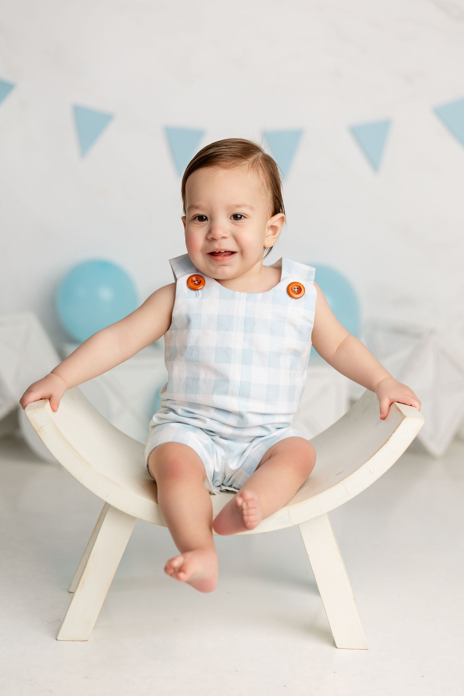 Infant wearing light blue romper sits happily in wooden prop chair with blue party decorations behind.