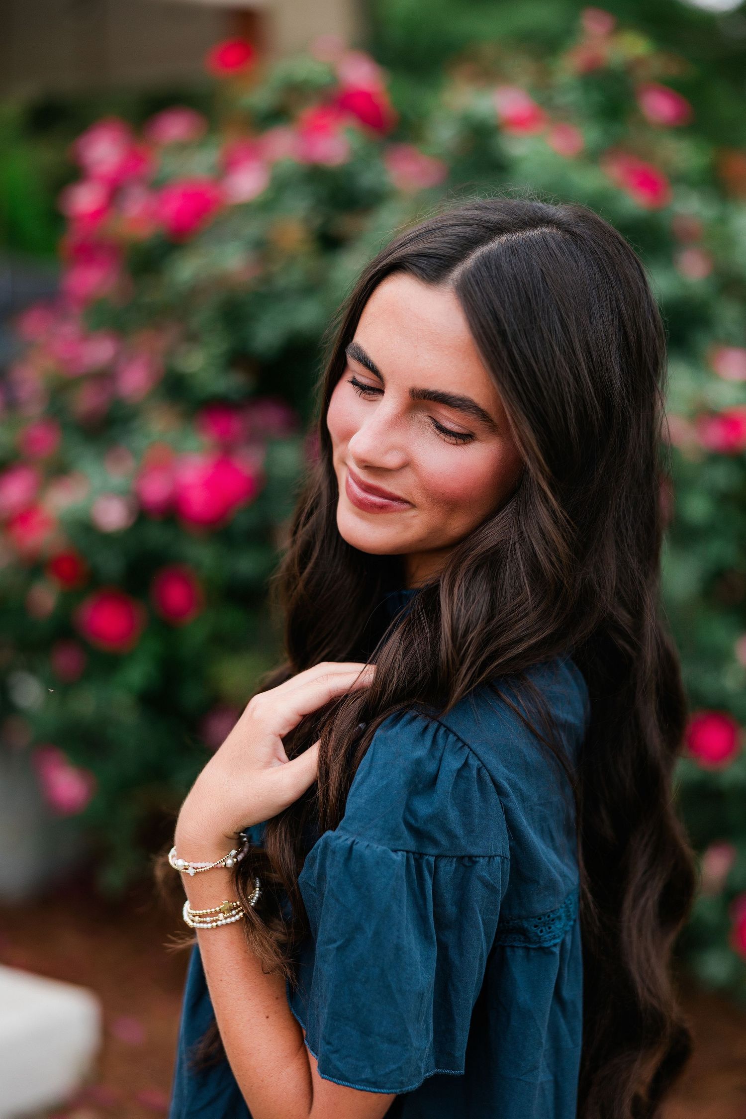 Young woman with long dark hair in teal dress smiling outdoors near blooming pink rose bushes.