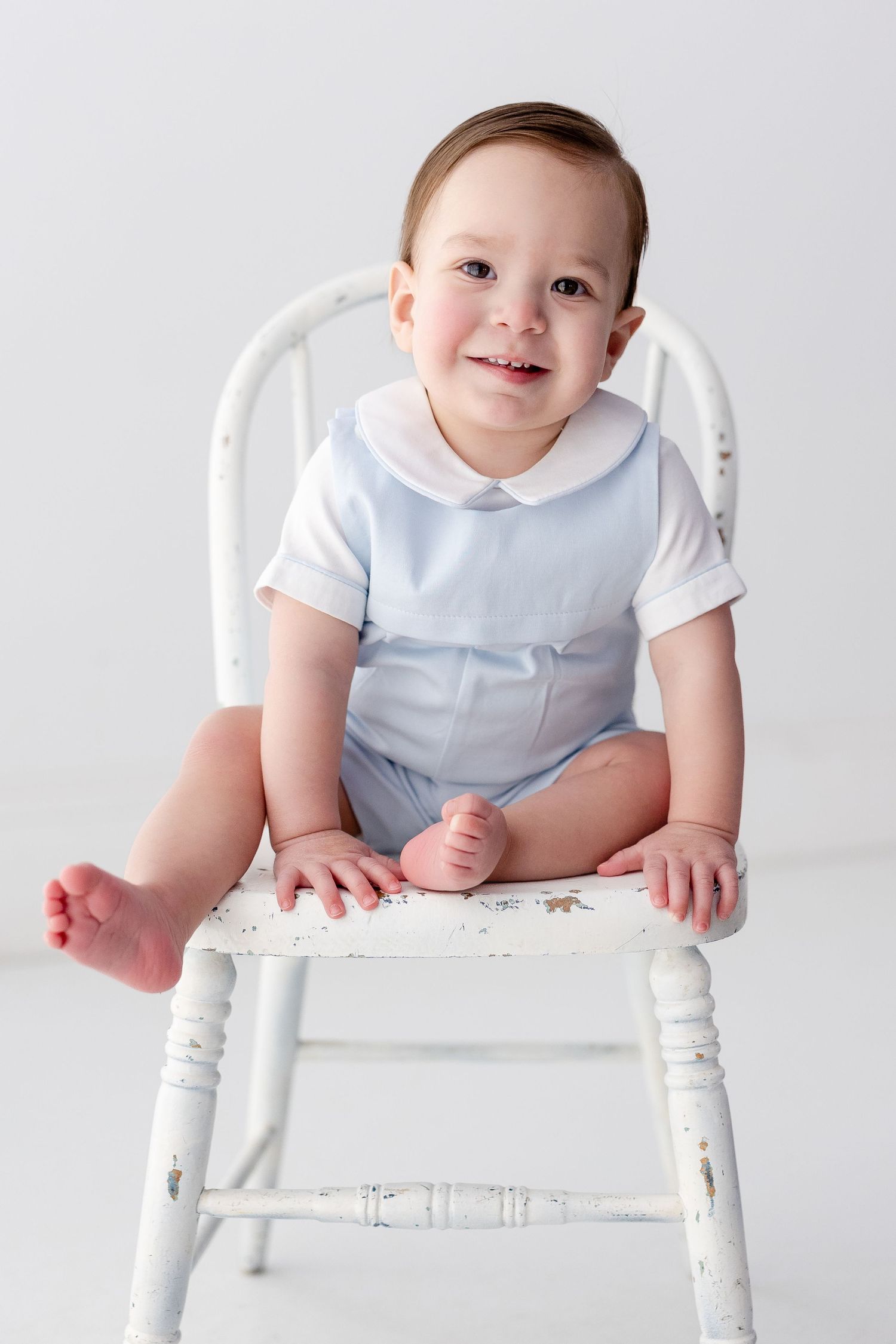 A toddler in a light blue and white outfit sits on a vintage white wooden chair against a white background.