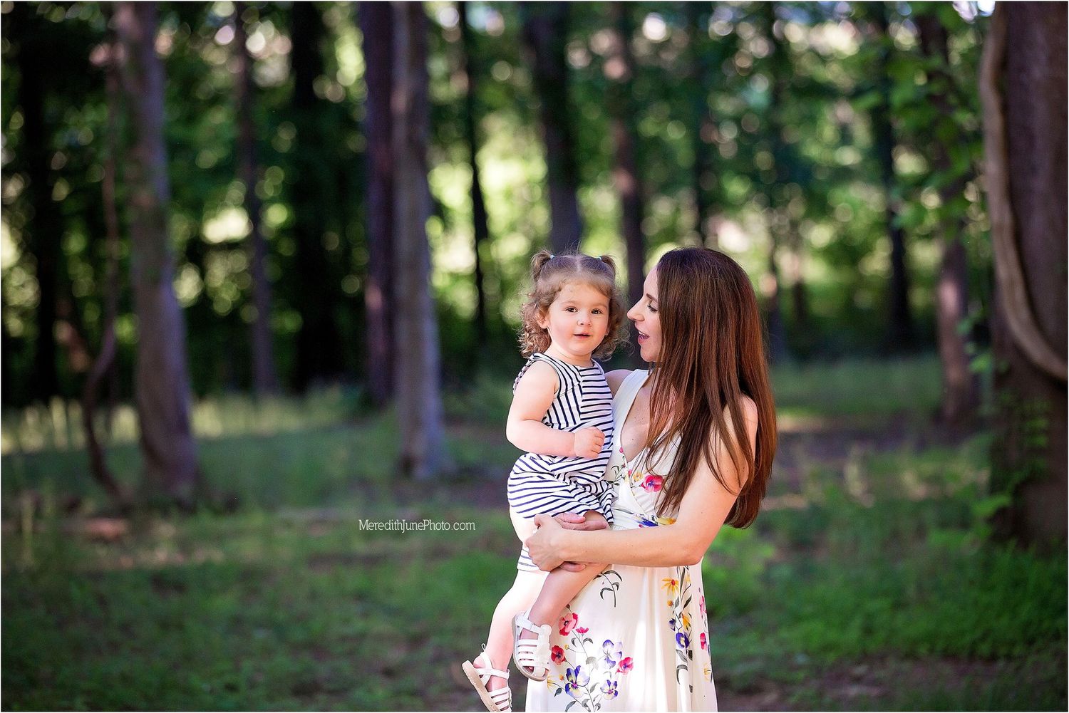 A woman in a floral dress holds a child while walking through a sunlit forest path during golden hour.