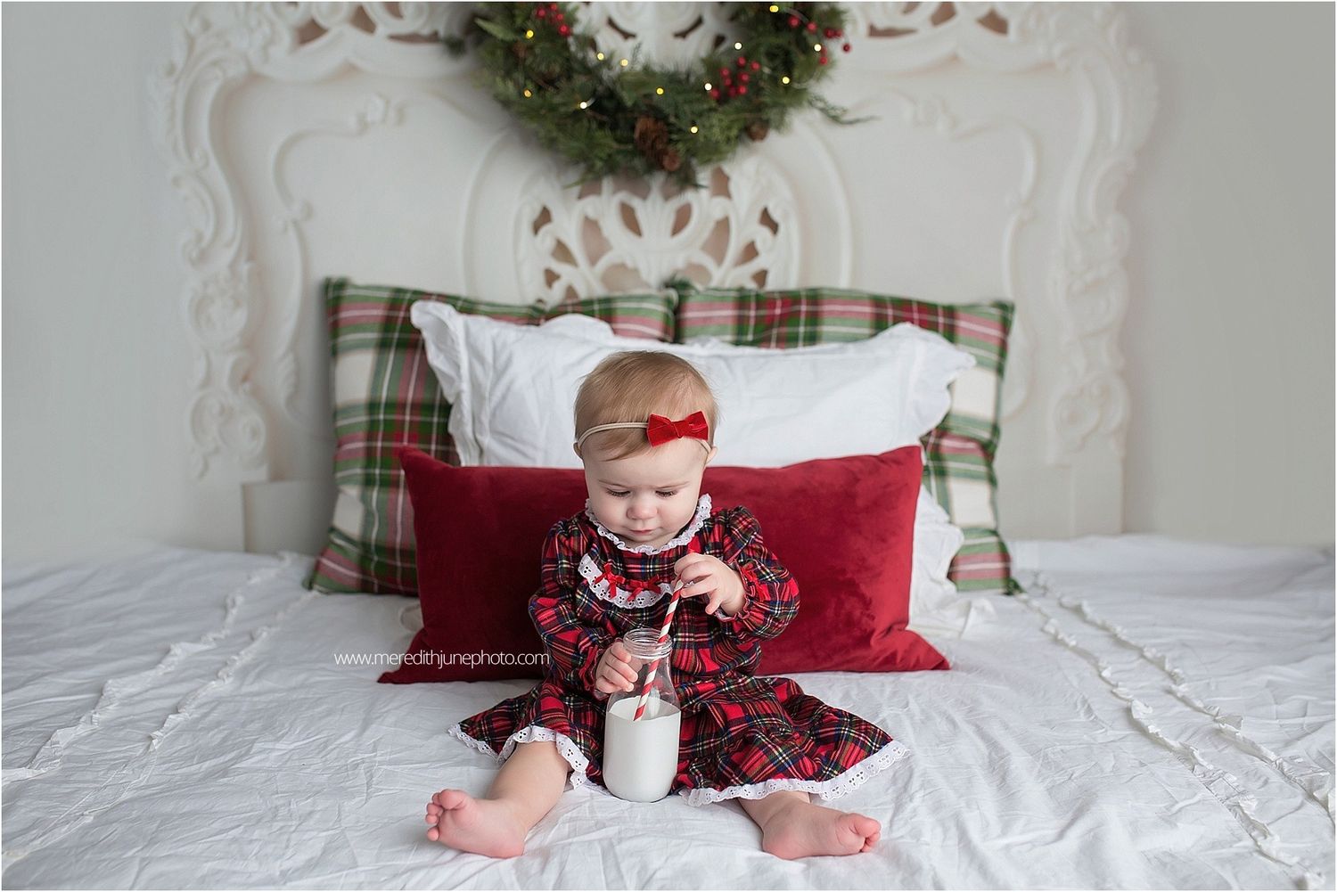 Child sitting on white bed with Christmas wreath and plaid decor in background.