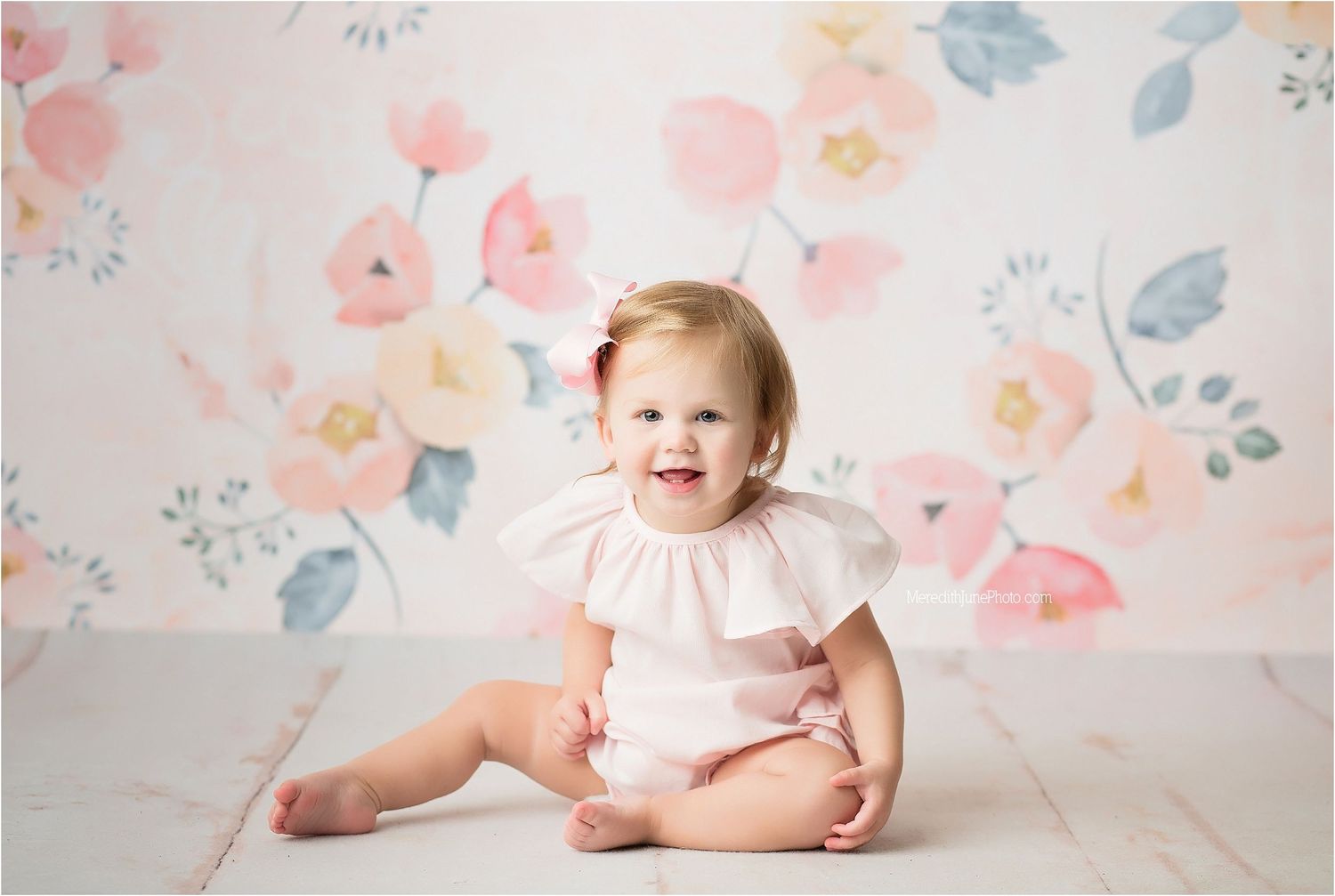 Baby in white ruffled outfit sitting on floor with floral wallpaper background.