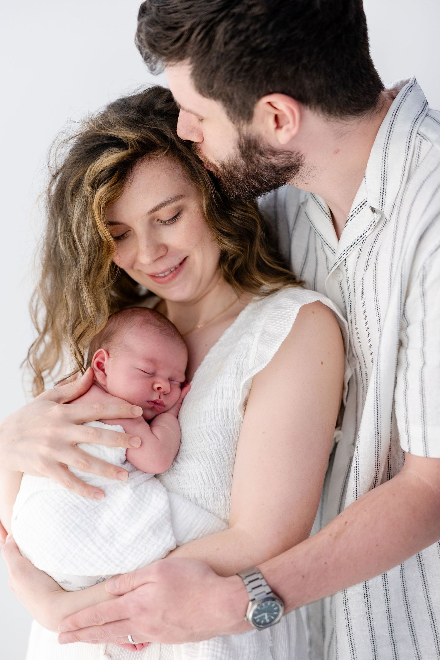 Happy young parents holding their sleeping newborn baby wrapped in white swaddle in a studio setting.