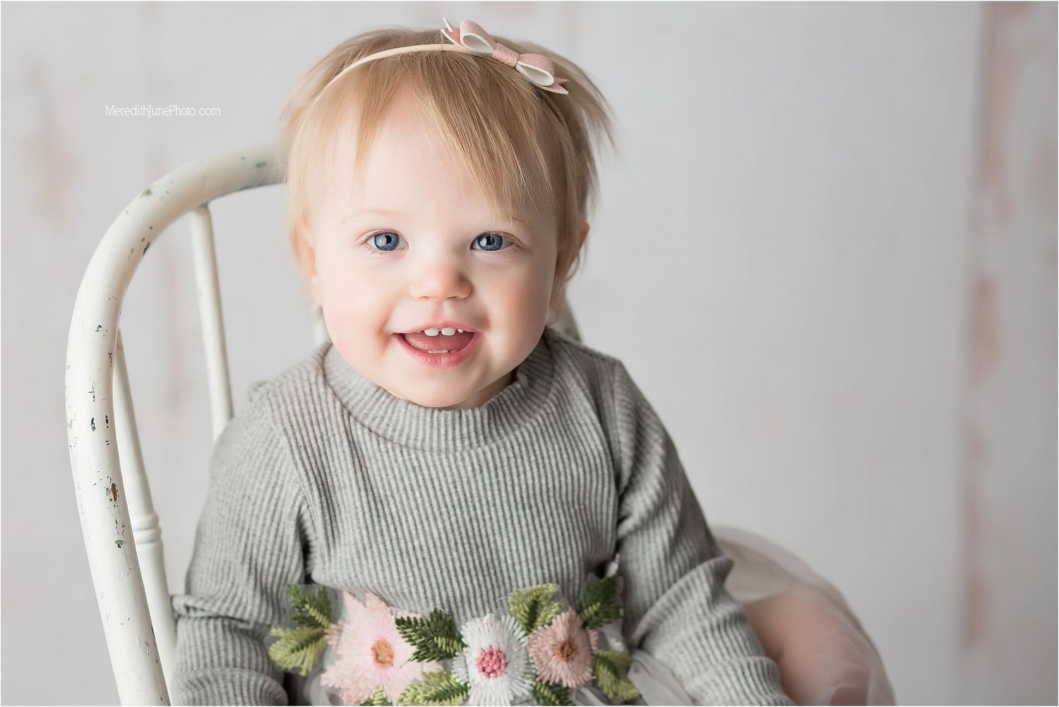 Toddler in gray sweater sits in white wooden chair with pink and white flowers.